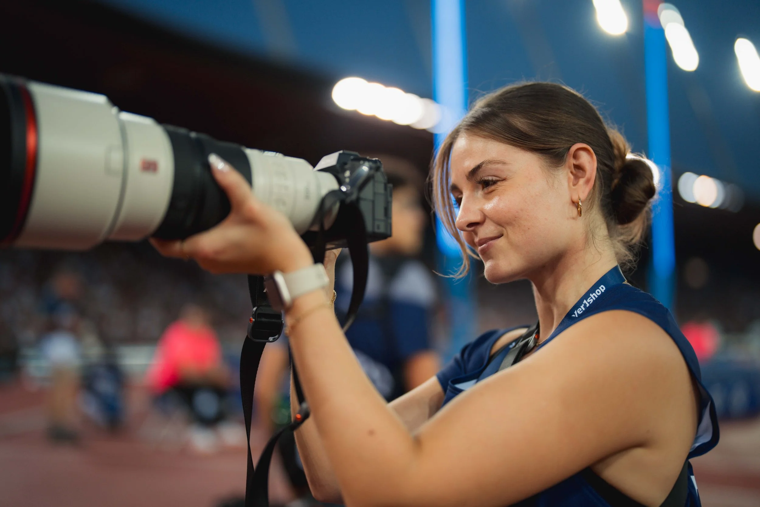 A woman with brown hair in a ponytail holding a professional camera on a tripod indoors, smiling, with blurred lights and people in the background.