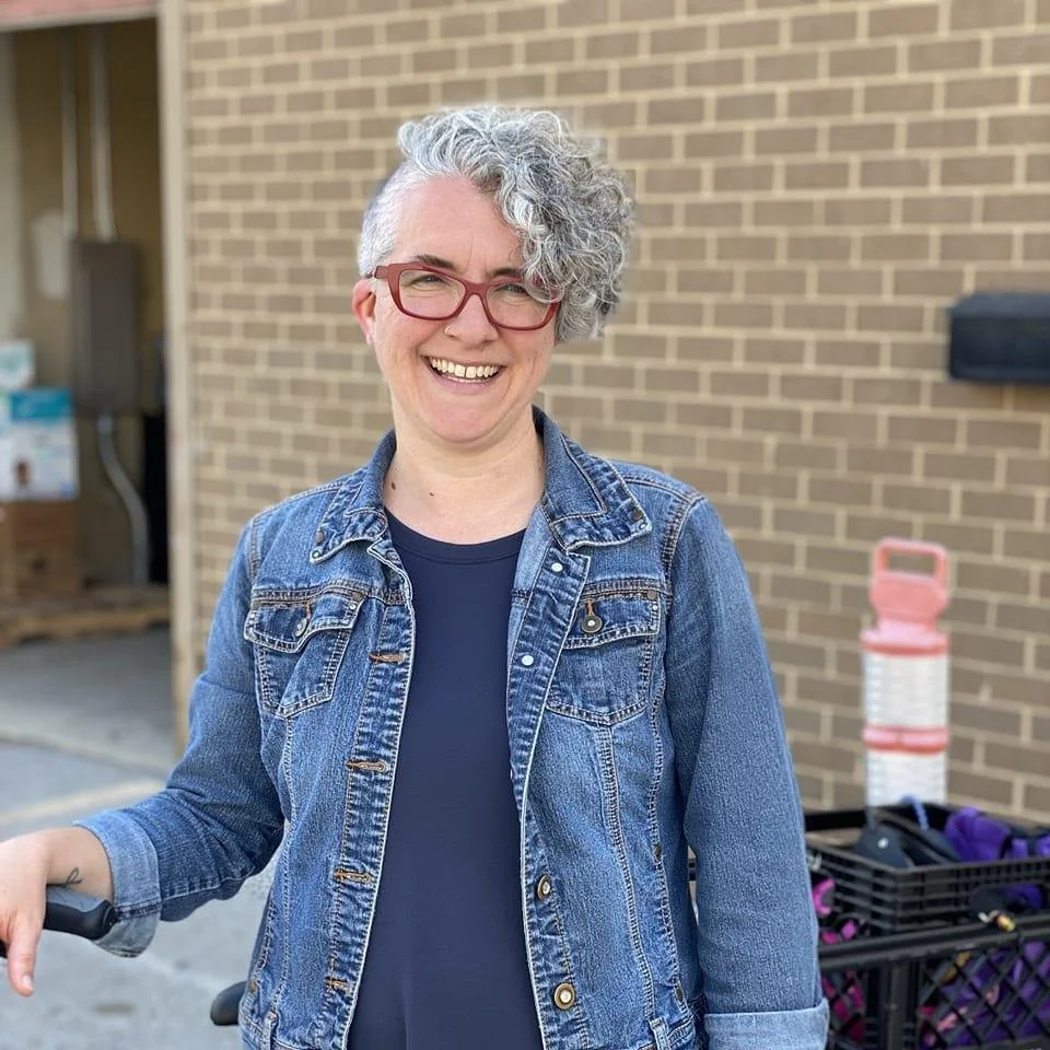 Rebecca Kolowe: A woman with curly gray hair and glasses smiling, wearing a denim jacket and standing outdoors in front of a brick wall.