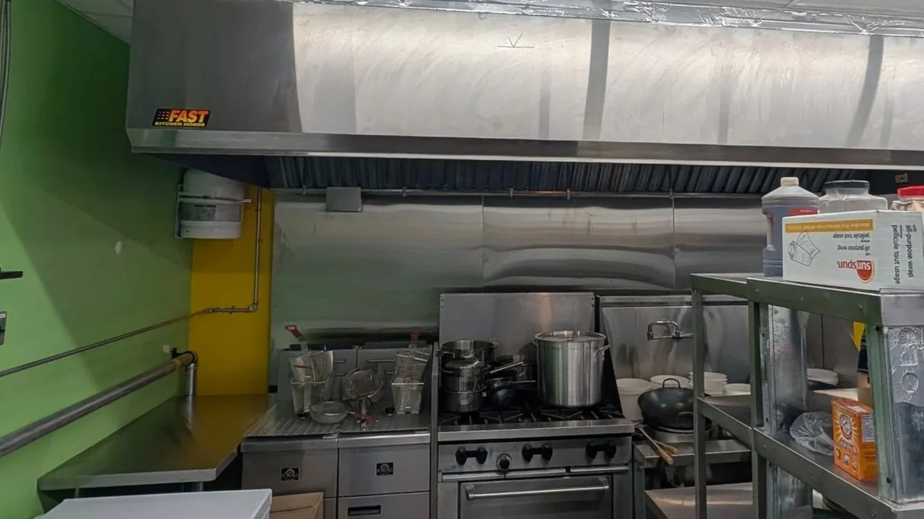 Commercial kitchen with stainless steel appliances, pots on stove, utensils, and shelves with containers, against green and yellow walls.