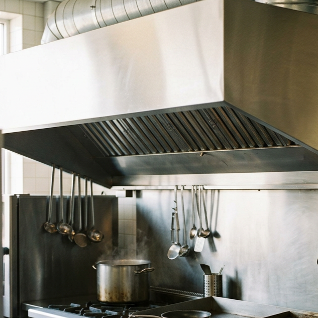 Commercial kitchen with a large stainless steel exhaust hood, hanging ladles and utensils, a pot on the stove with steam, and various kitchen tools.