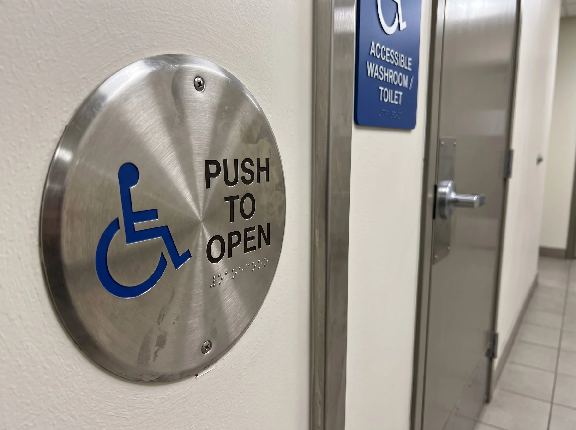 Close-up of a silver wheelchair-accessible door push plate with text 'PUSH TO OPEN' mounted on a cream-colored wall, with a blue sign for accessible washroom/toilet in the background.