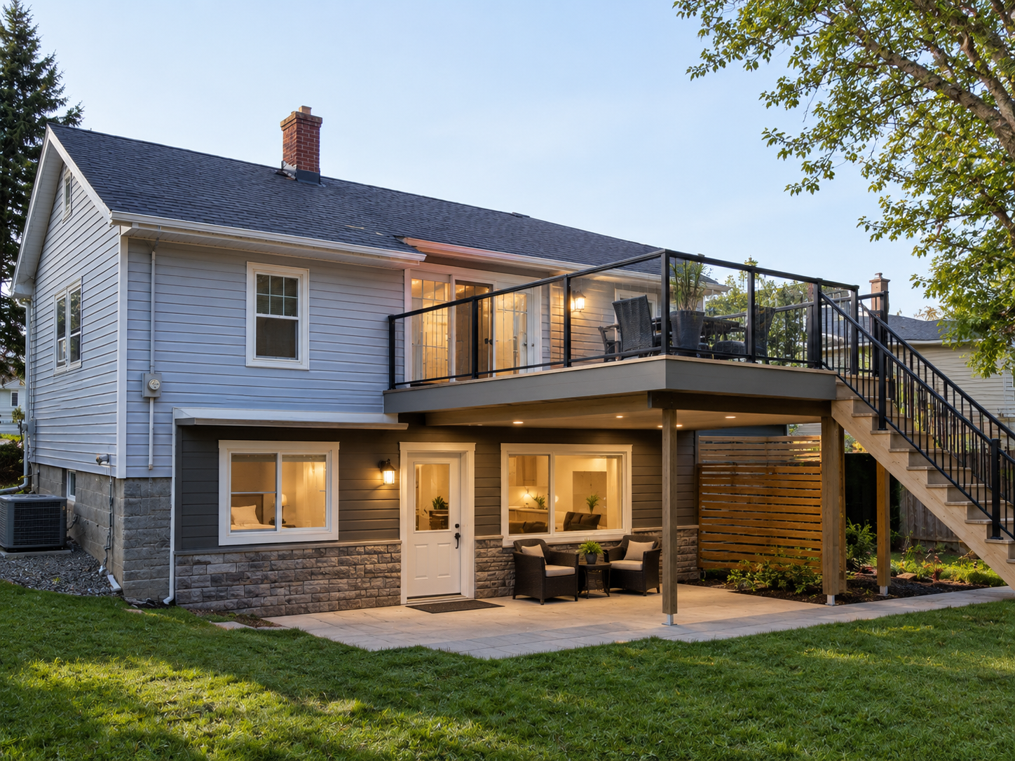 A two-story house with a basement, outdoor patio, and deck at sunset. The house has light blue siding, a stone foundation, and windows with white frames. The upper deck has a sitting area with chairs, a table, and plants, connected to the backyard by a staircase with black railings. The backyard has grass and a small garden bed, and screened porch doors lead into the house. The lighting is warm inside, visible through the windows and doors.