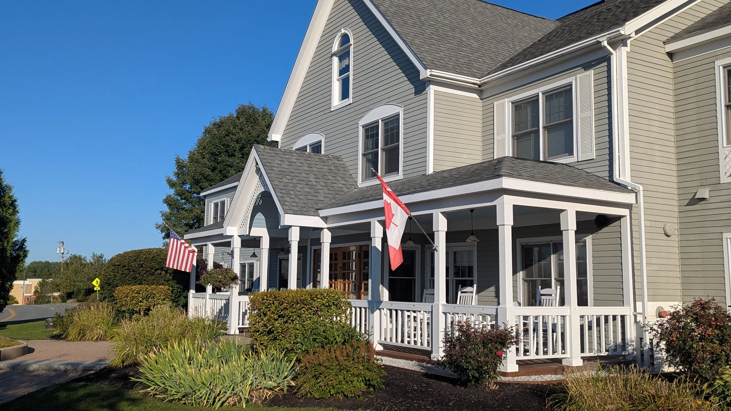 A large two-story house with a porch, light gray siding, white trim, and American flags hanging from the porch. The house has multiple windows and a steep roof, set against a clear blue sky.