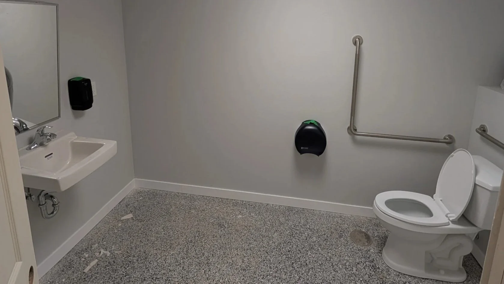 Empty accessible restroom with a toilet, a small sink, grab bars, and a black soap or sanitizer dispenser on the wall.