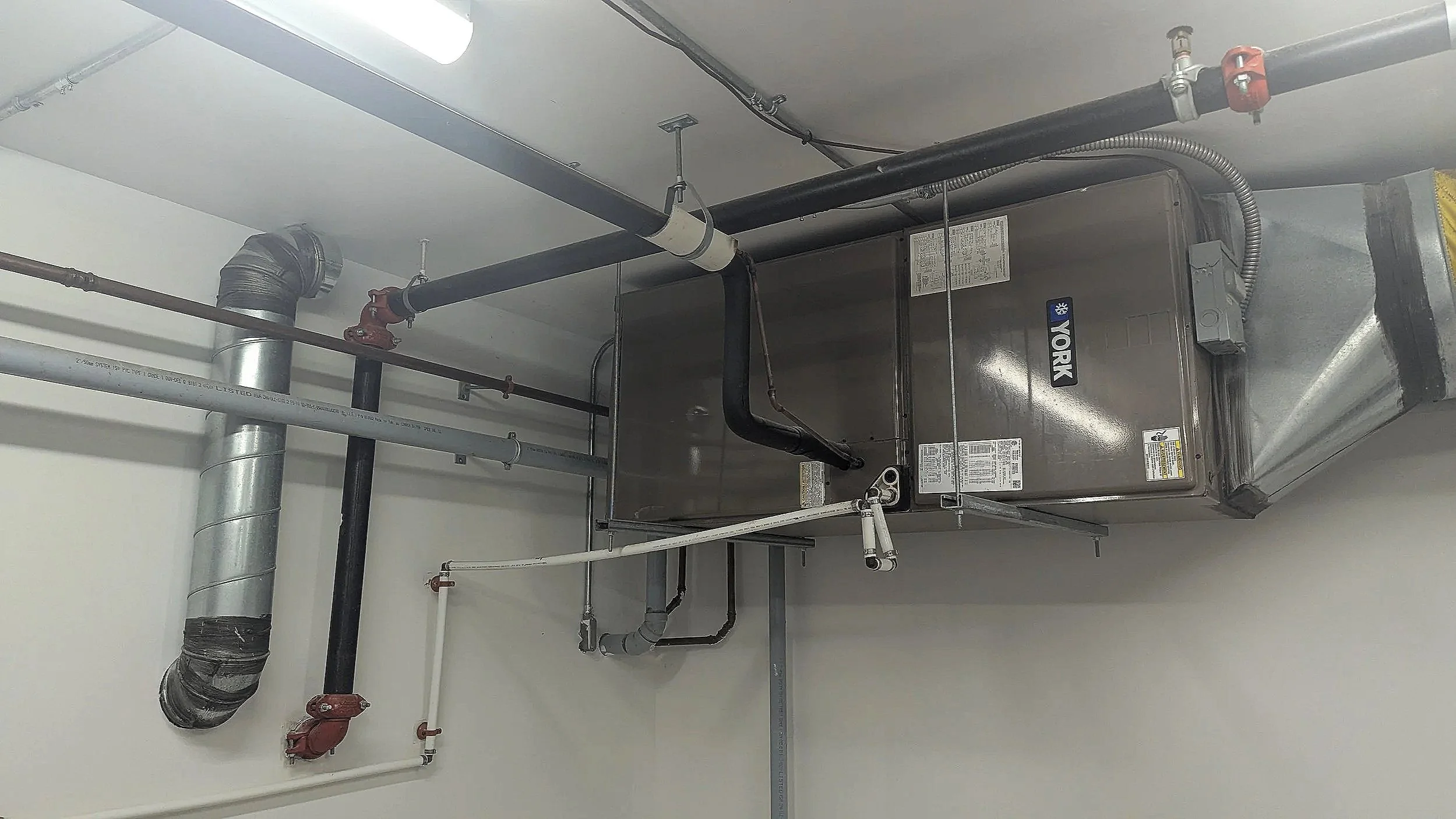 A commercial HVAC air conditioning unit installed on a ceiling with black and silver pipes, some with red valves, and white and black PVC pipes in a room with a white ceiling and wall.