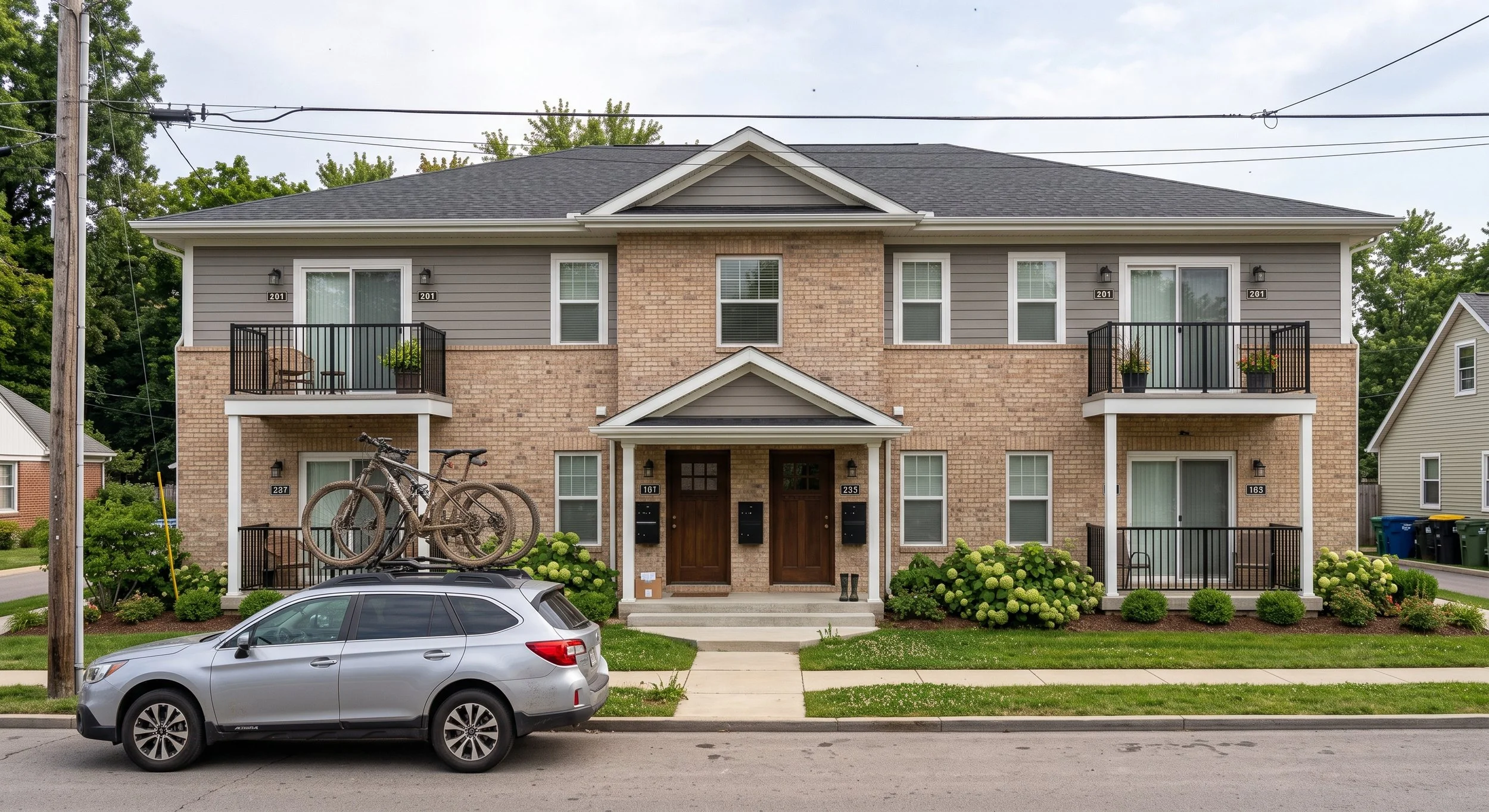 A multi-story residential building with brick and siding exterior, featuring small balconies with potted plants, a front porch, and a parking space in front where a silver car and a bike rack with bikes are parked.