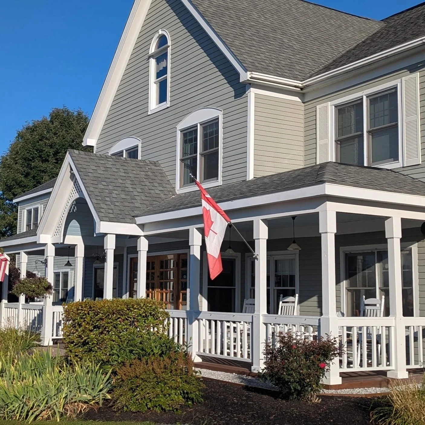 A multi-story house painted in light gray with white trim, a porch with white railings, and a Canadian flag flying in front. There is a garden with bushes and plants in the foreground.