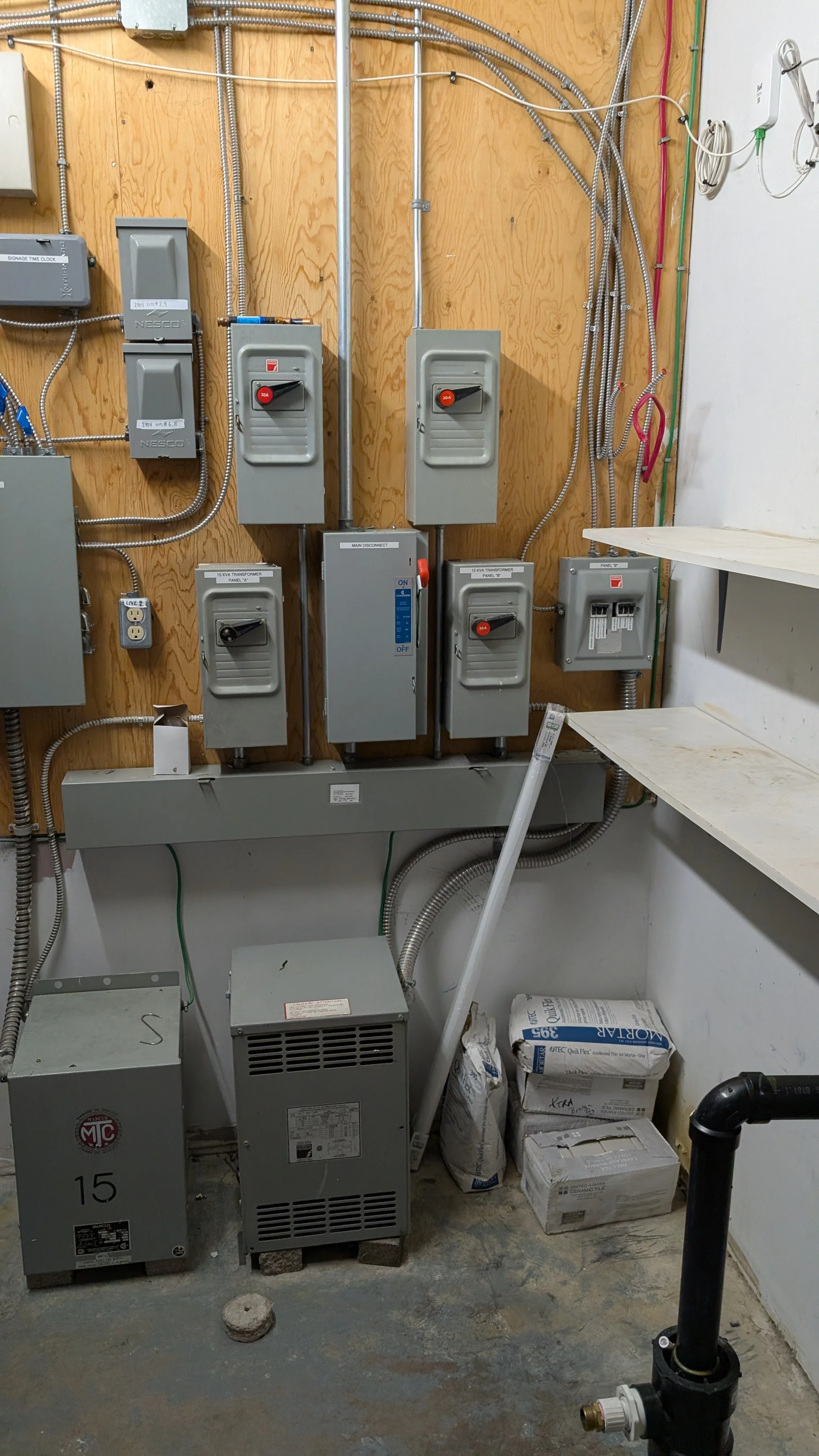 Electrical panels, transformers, and wiring mounted on a wooden wall in a utility room, with some bags of material on the floor and a pipe in the foreground.