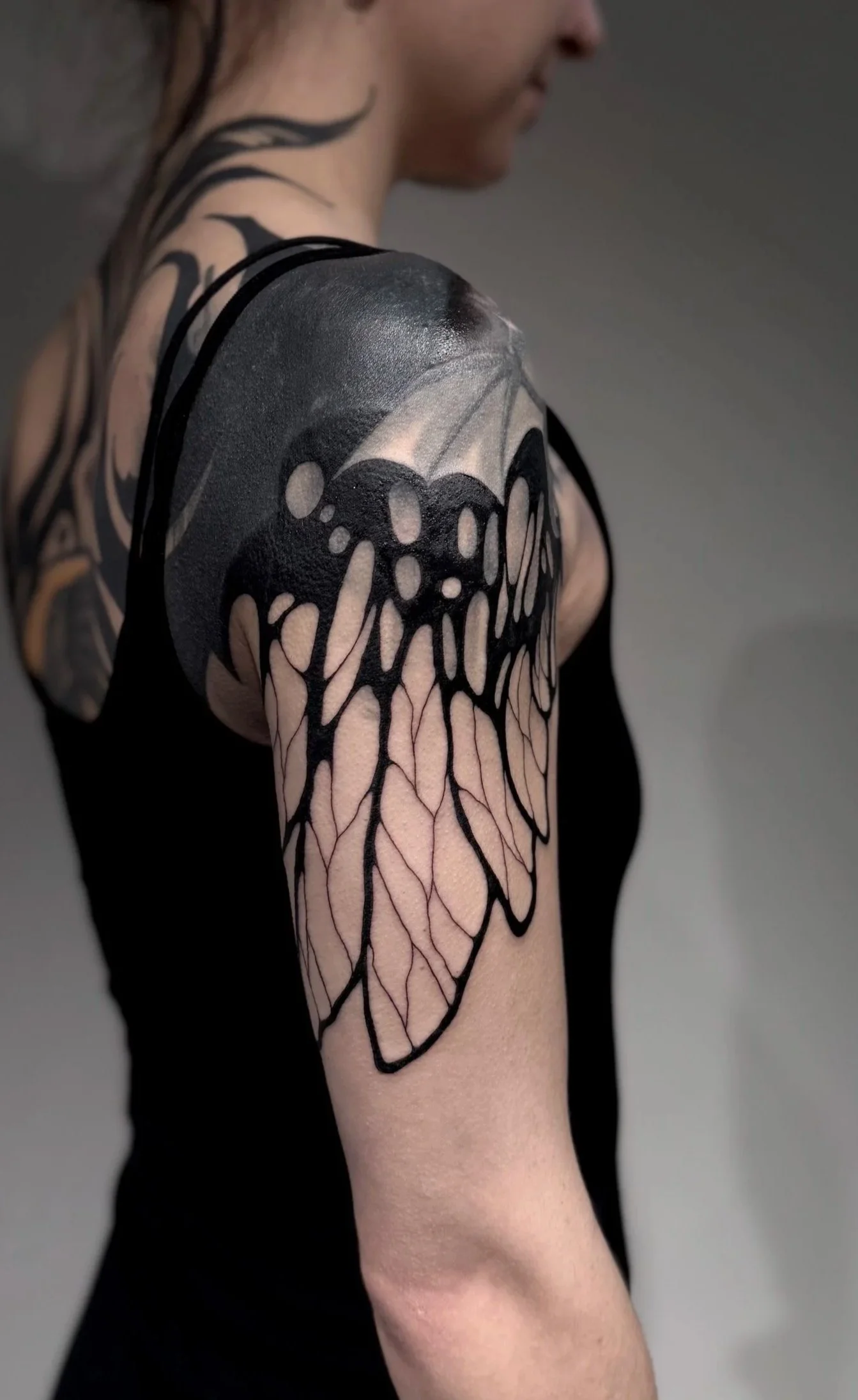 Nahaufnahme eines menschlichen Schulter- und Oberarms mit schwarzem Tattoo, das ein Flügelmuster darstellt, gegen einen grauen Hintergrund.