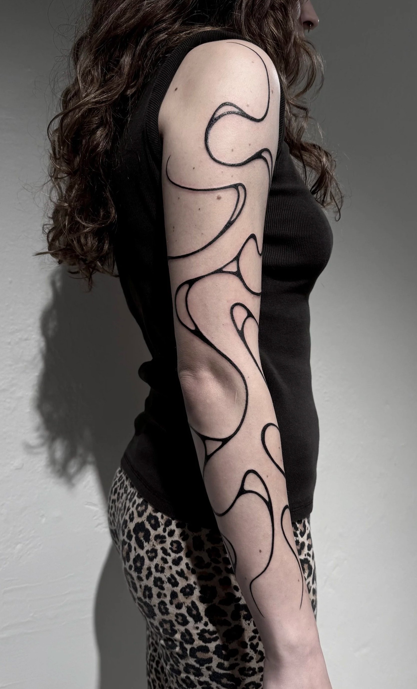 Junge Frau mit langen, lockigen braunen Haaren, schwarzem ärmellosem Top und Leo-Print-Hose, zeigt Silhouetten-Tattoo auf ihrem linken Arm gegen grauen Hintergrund.