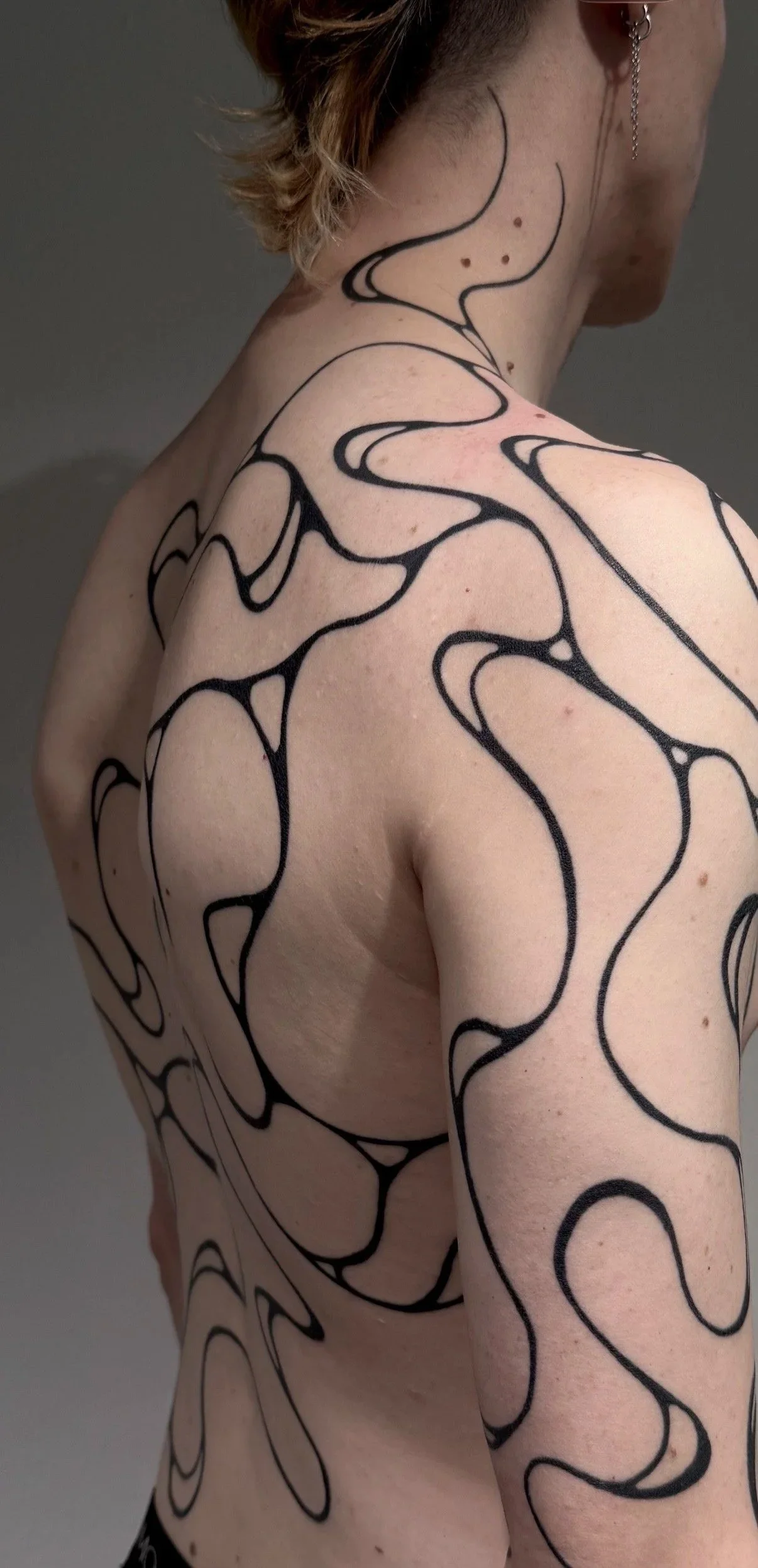 Nahaufnahme eines Menschen mit Tattoo-Designs aus schwarzen, geschwungenen Linien auf Schulter, Arm, Brust und Rücken.