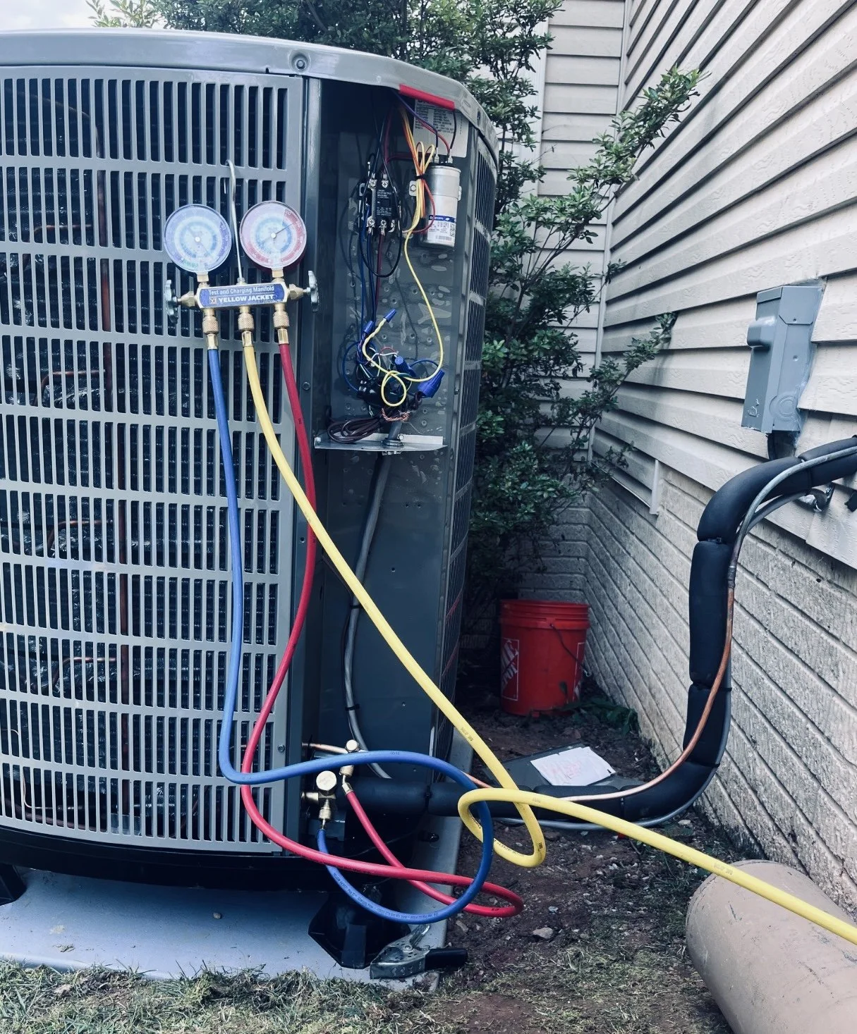 An outdoor air conditioning unit connected with yellow, blue, and red refrigeration hoses, with gauges attached, next to a house with siding, a bush, a red bucket, and an electrical box on the wall.