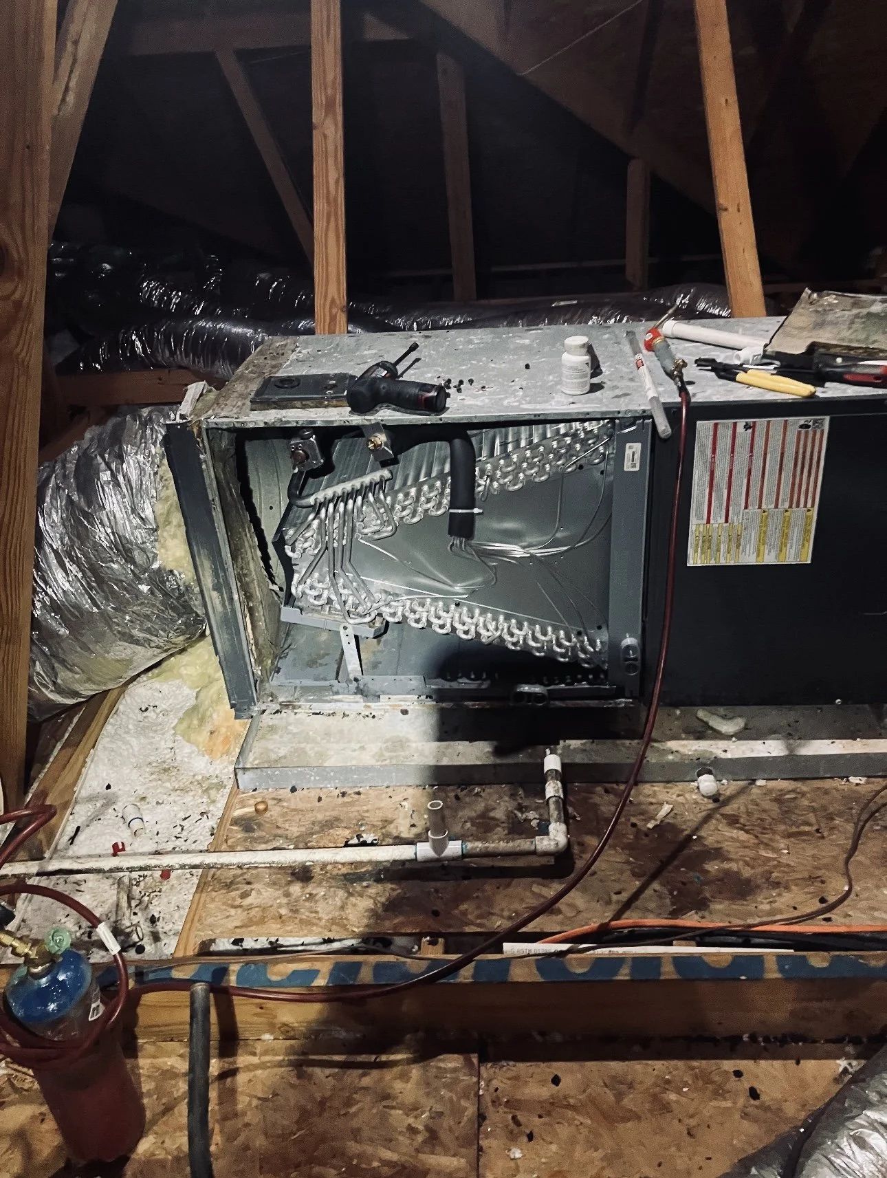 Open dishwasher inside an attic with tools and construction materials on top, exposed wooden beams, and insulation on the walls and floor.
