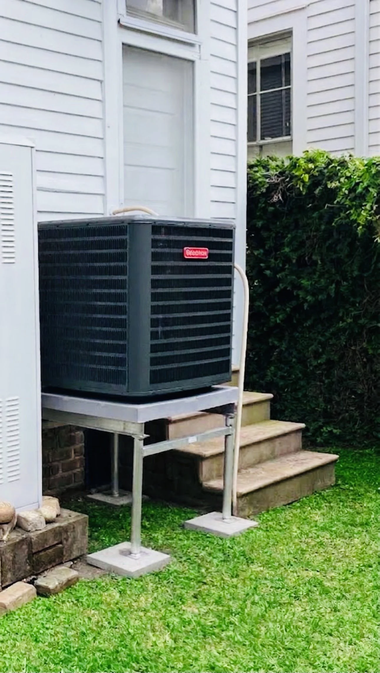 An outdoor air conditioning unit on a platform beside a house, with a small staircase and green grass in the foreground.