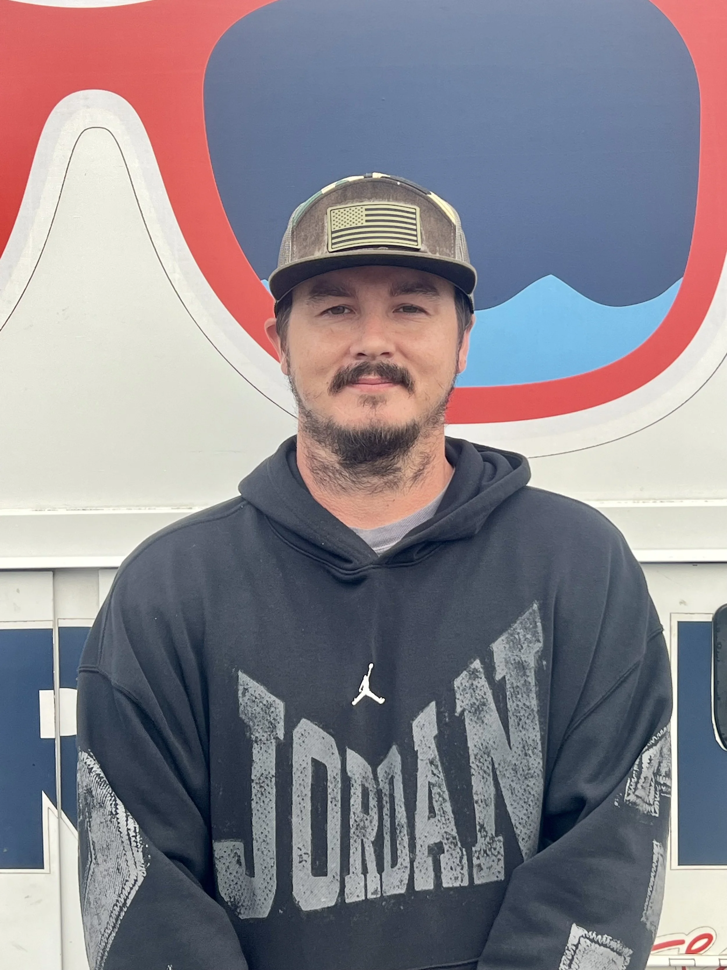 A man with a beard and mustache wearing a camouflage cap with an American flag patch and a black hoodie, standing in front of a colorful background with red, white, and blue elements.