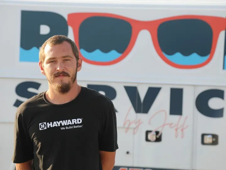 A young man with short hair and a beard, wearing a black T-shirt with the Hayward logo, standing in front of a sign that says 'Pool Service' and features a large image of sunglasses with a wave pattern in the lenses.