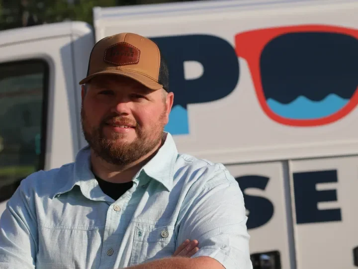 A man with a beard and mustache in a light blue button-up shirt and a tan trucker hat, standing with arms crossed in front of a sign with the word 'POLICE' and a logo showing a blue and red shape.