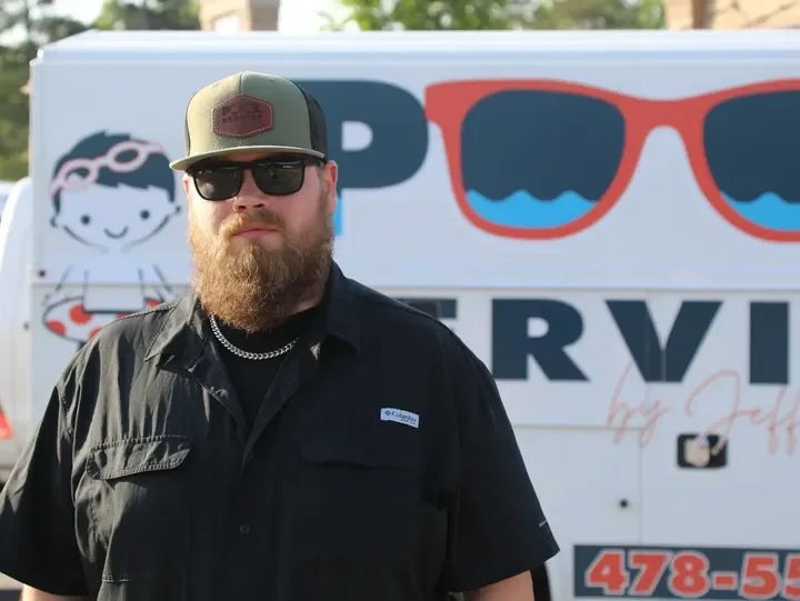 Man with a beard wearing sunglasses, a black shirt, a baseball cap, and a chain necklace, standing in front of a vehicle with sunglasses printed on it.