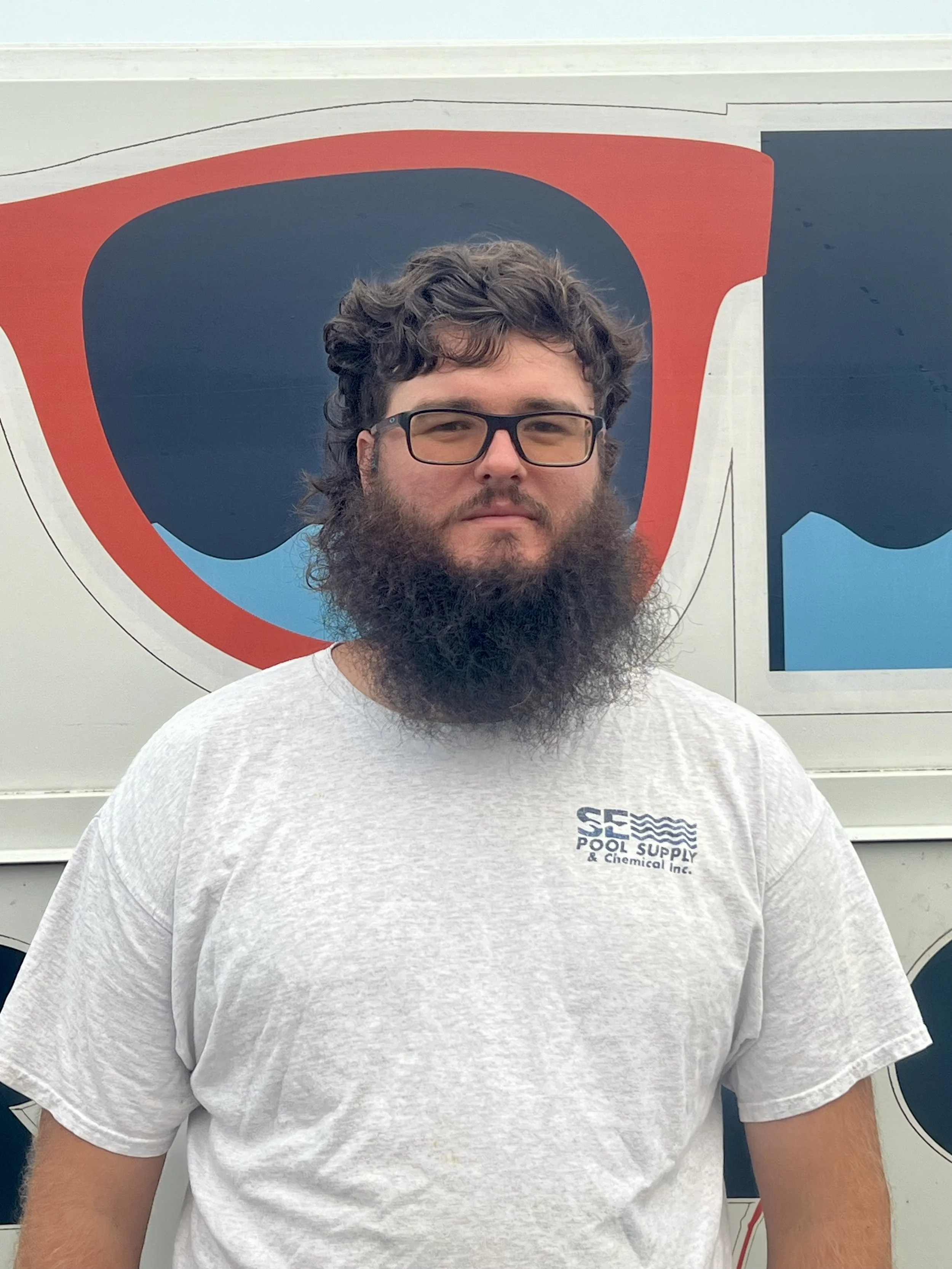 A man with curly dark hair, glasses, and a full beard wearing a white T-shirt with a logo that reads 'SFS Pool Supply & Chemical Inc.' standing in front of a sign with a large red, black, and blue design.