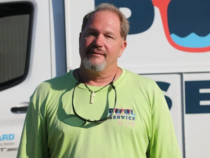 A man with short, light brown hair and a beard, wearing a neon green long-sleeve shirt with a logo and text, standing outdoors in front of a white vehicle with a logo.