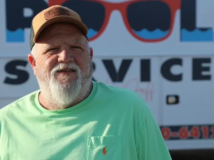 A man with a gray beard wearing a tan cap and a light green T-shirt with a small orange logo on the pocket, standing outdoors in front of a vehicle with bold text and sunglasses on the sign.