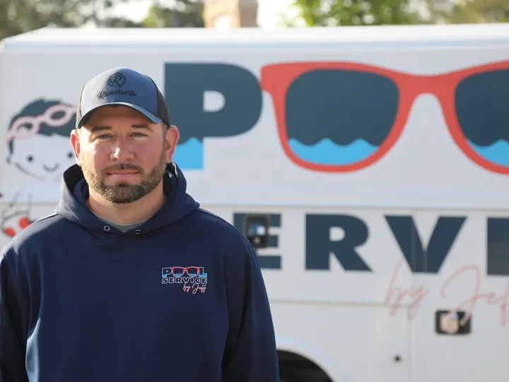 A man wearing a baseball cap and a hoodie stands outdoors in front of a service van with sunglasses logo.