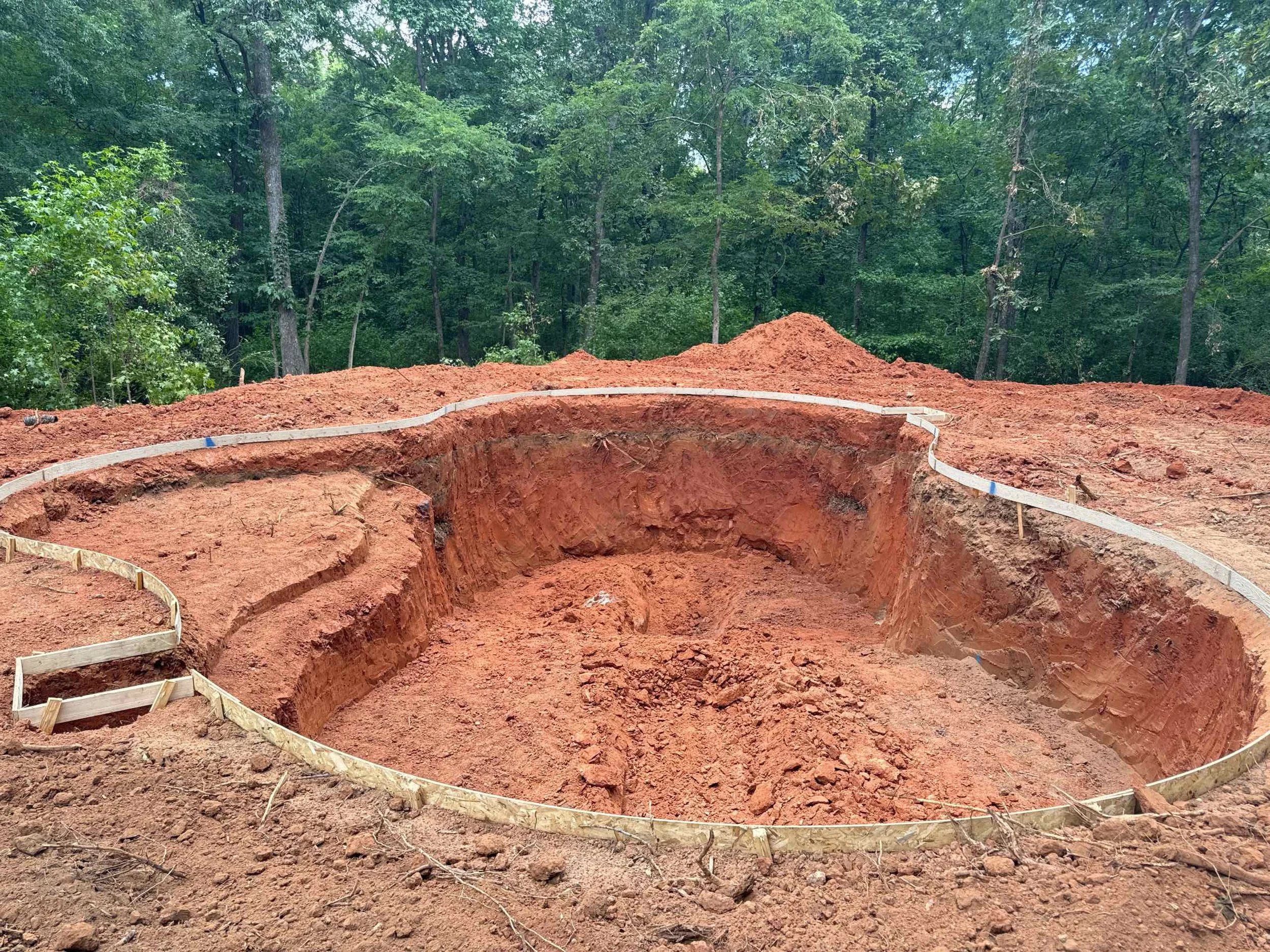 A construction site with an excavated circular hole in the ground, surrounded by red soil and a wooded area in the background.