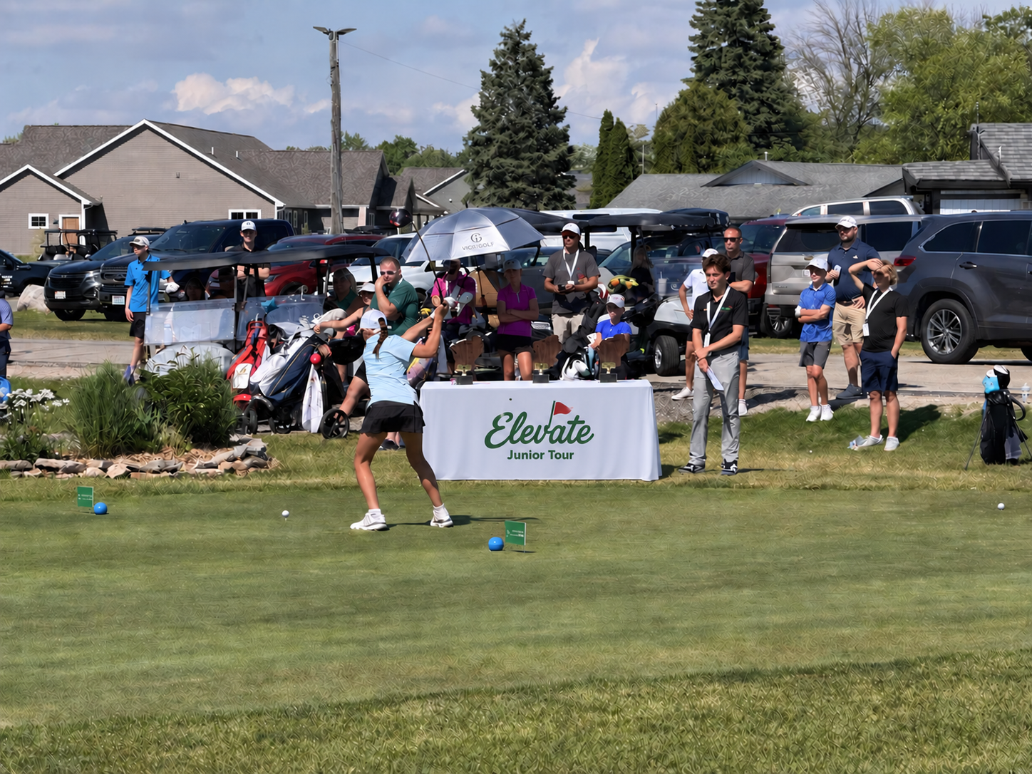 A junior golfer tees off during an Elevate Junior Tour tournament while players and families watch.