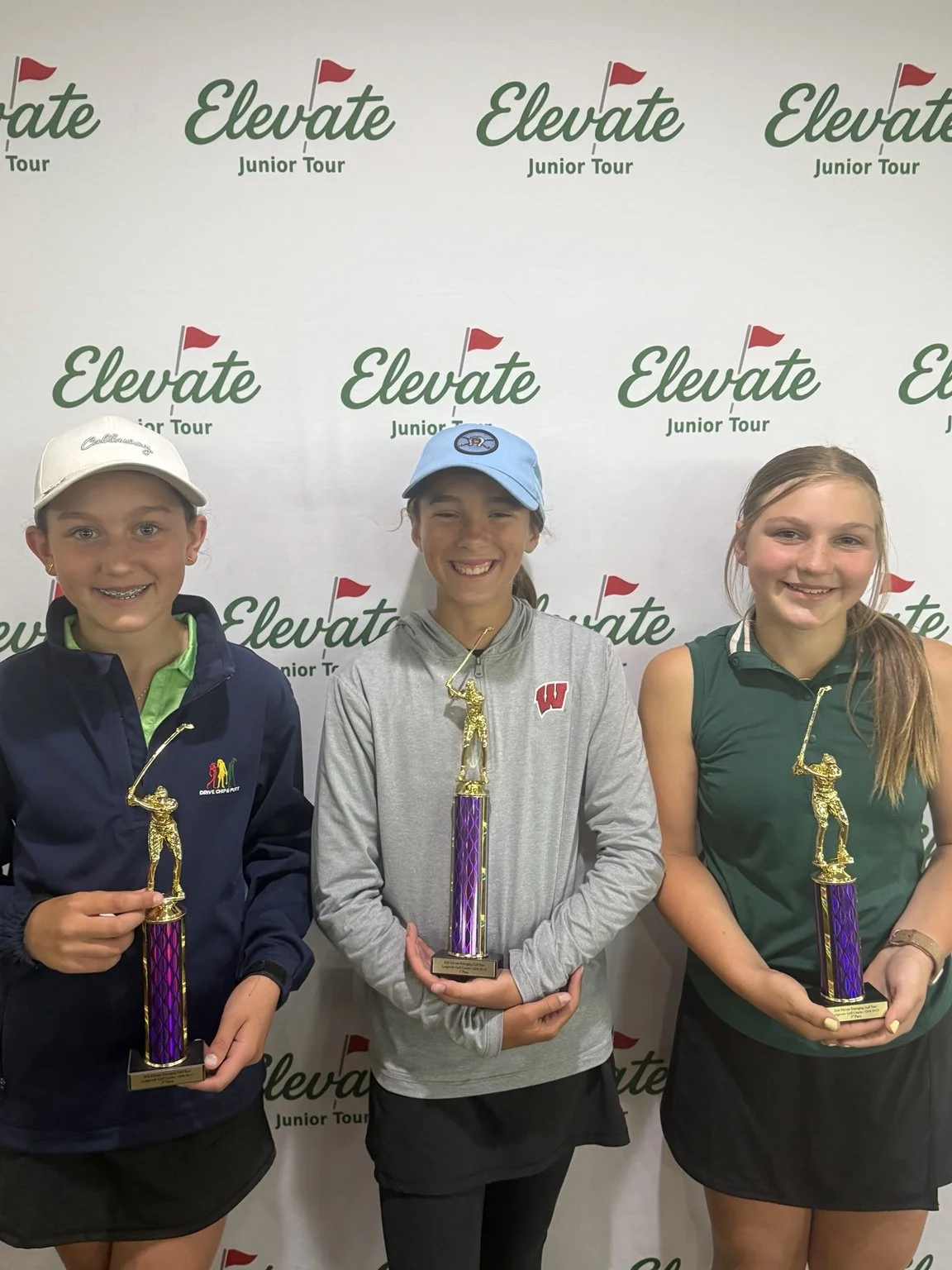 Three Emerging Tour girls hold purple trophies in front of the Elevate Junior Tour backdrop.