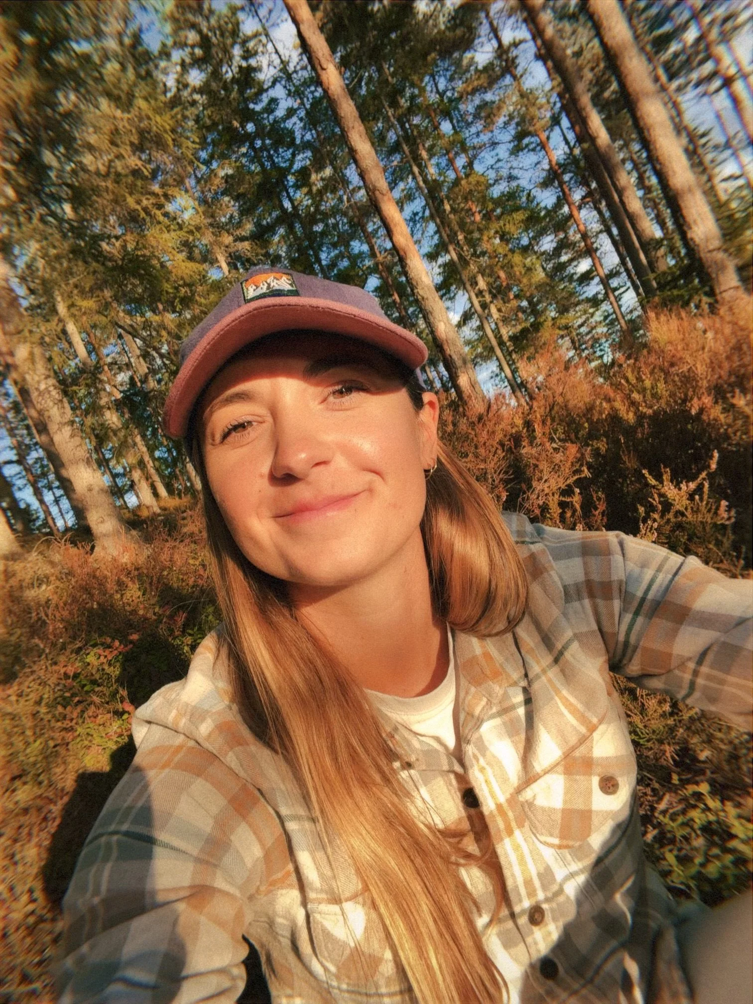 A young woman with long red hair smiling outdoors in a forest during sunset, wearing a plaid shirt and a baseball cap.