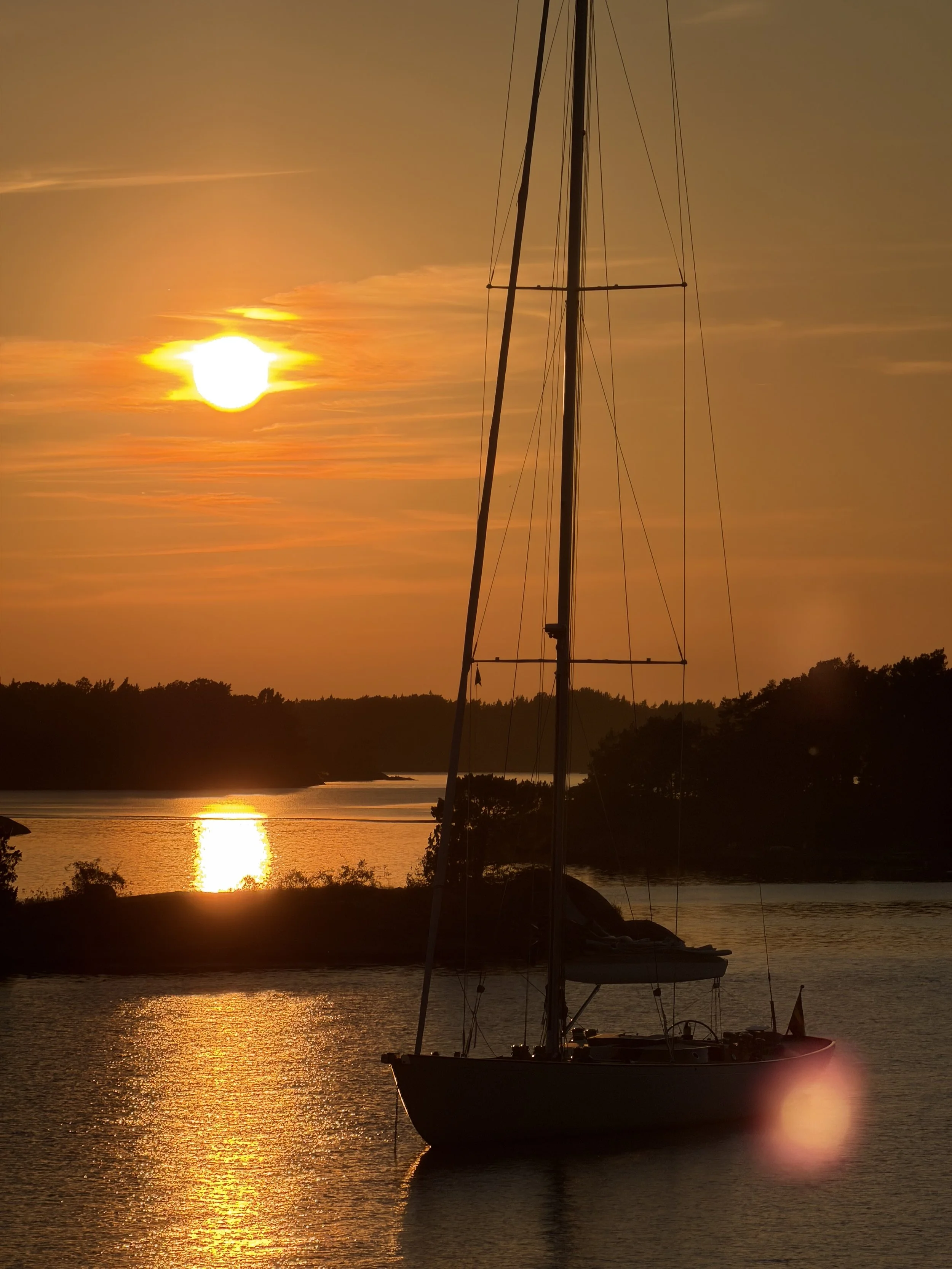 A sailboat docked in a calm body of water during a sunset, with the sun casting a golden glow across the sky and reflecting on the water's surface.
