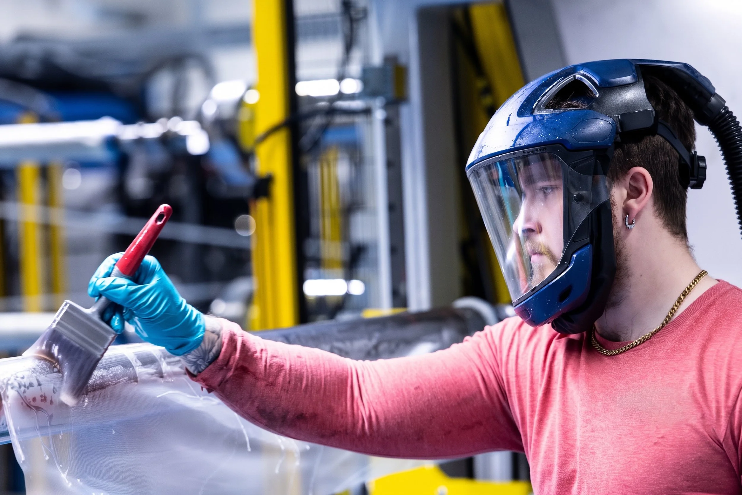 A person wearing a blue helmet with a clear face shield, a red shirt, blue gloves, and a gold chain, working with a piece of equipment in an industrial or manufacturing setting.