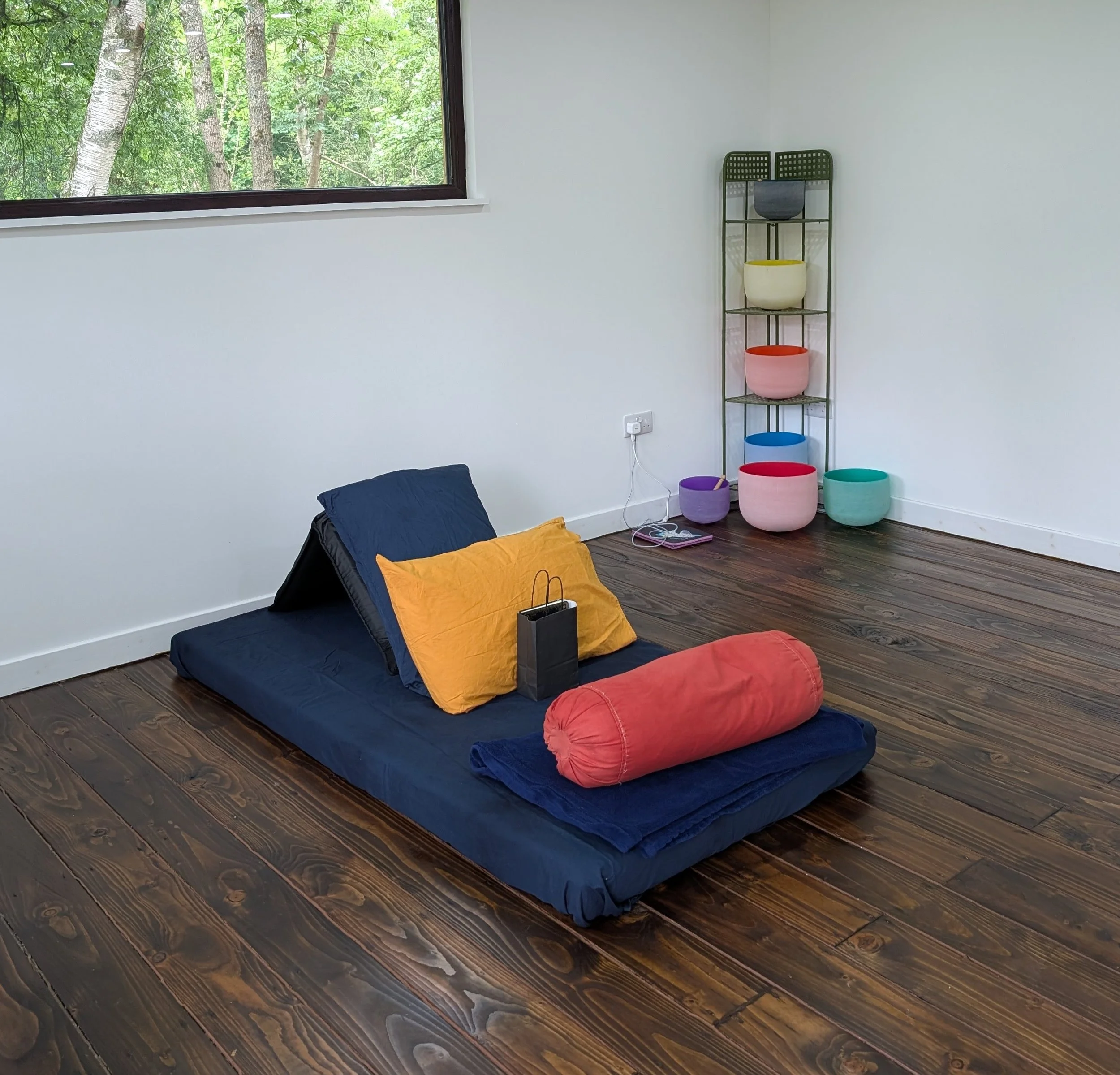 A room with wooden flooring, a large window showing green trees outside, and a black metal shelf holding colourful bowls. There are cushions and pillows on the floor.
