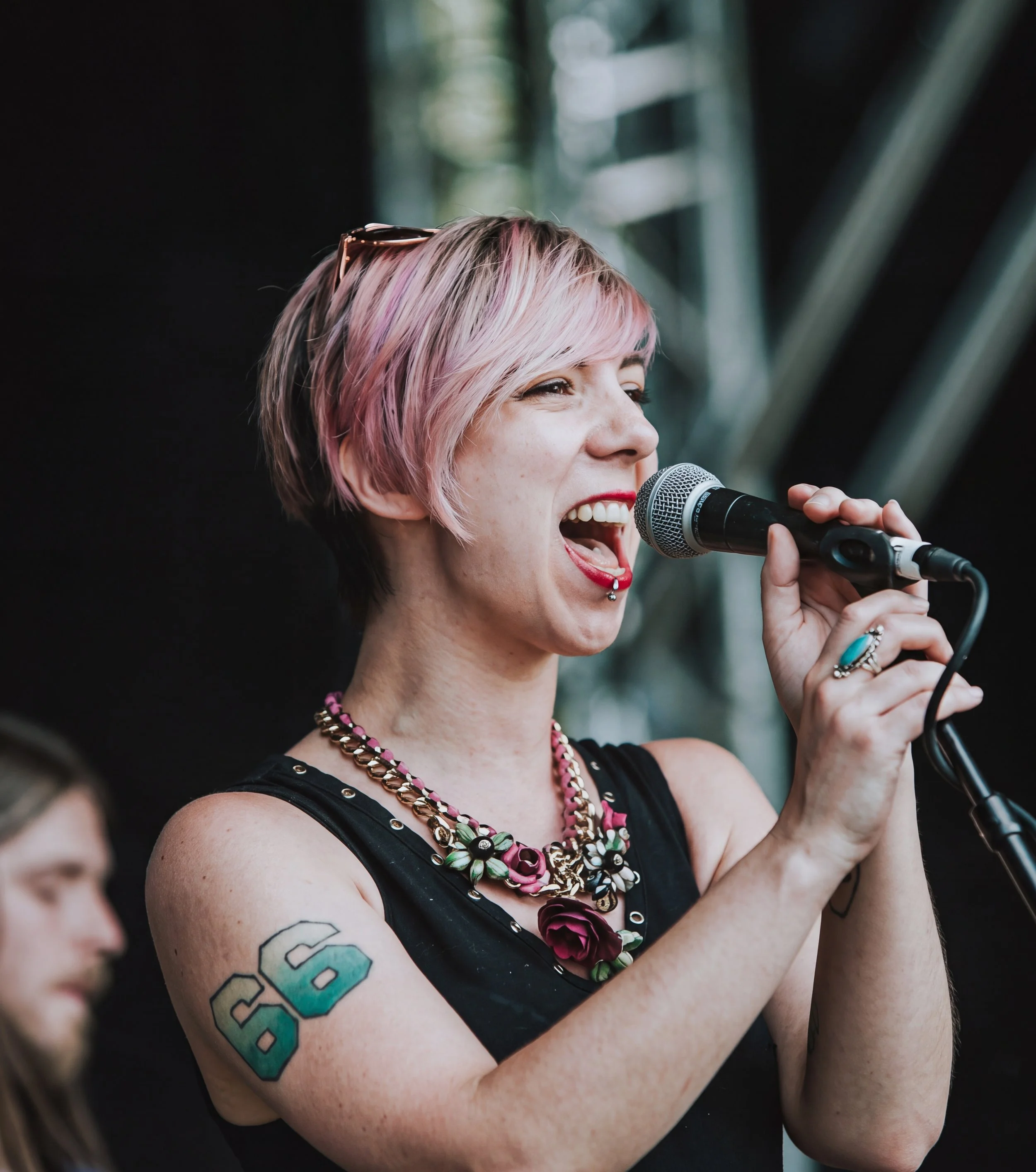 Mo Ford - a person with short pink and blonde hair singing into a microphone at an outdoor event, wearing a black tank top with metal studs and colourful necklaces