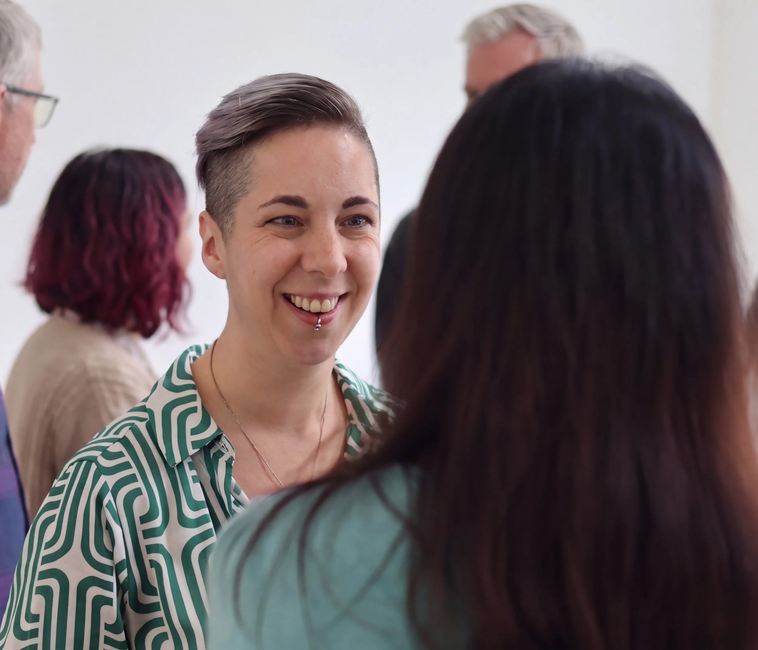 Mo Ford, smiling, with short hair and a lip piercing, engages with a group of people in a discussion or meeting.