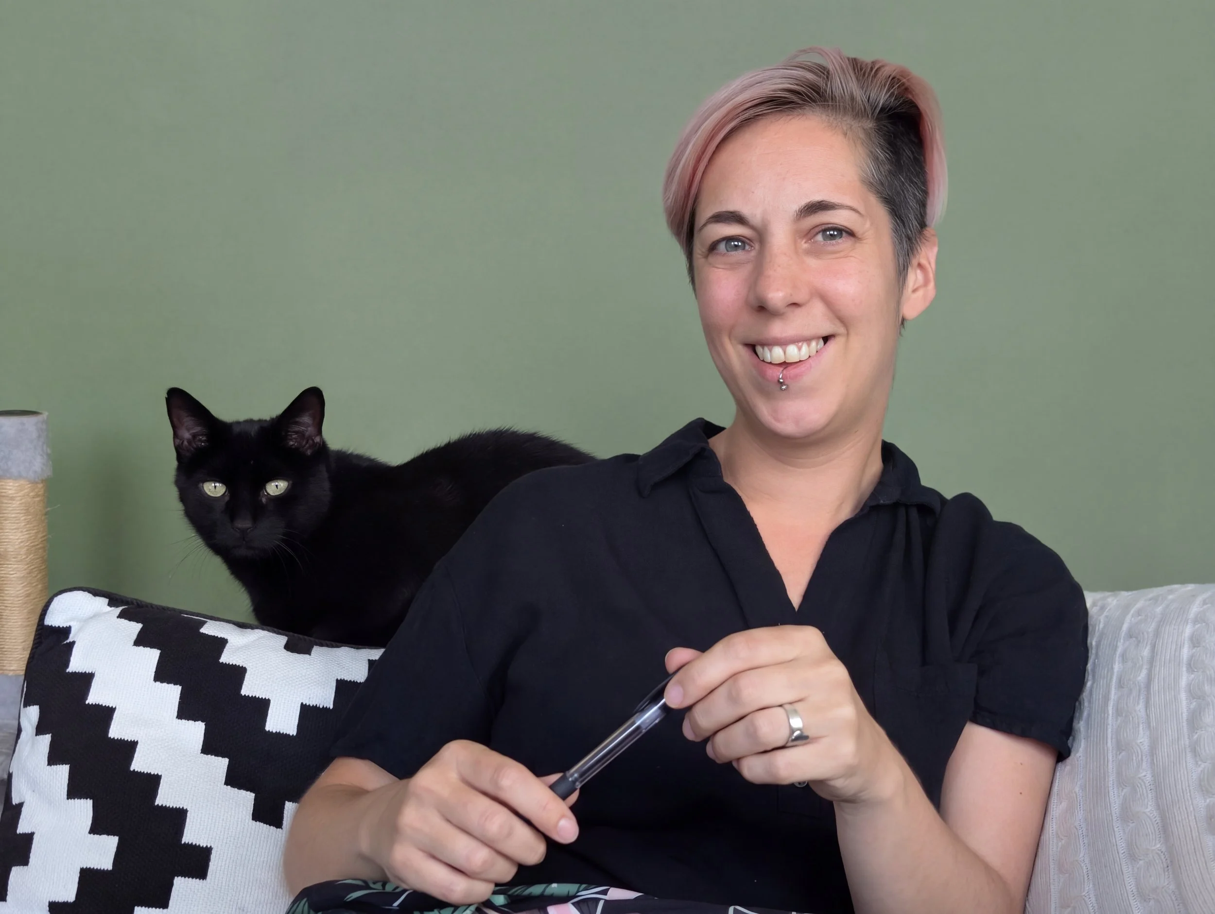 Mo Ford, a person with short pink and black hair, smiling, holding a pen, sitting on a sofa with a black and white checkered pillow and a gray cushion, next to a black cat, against a green background.