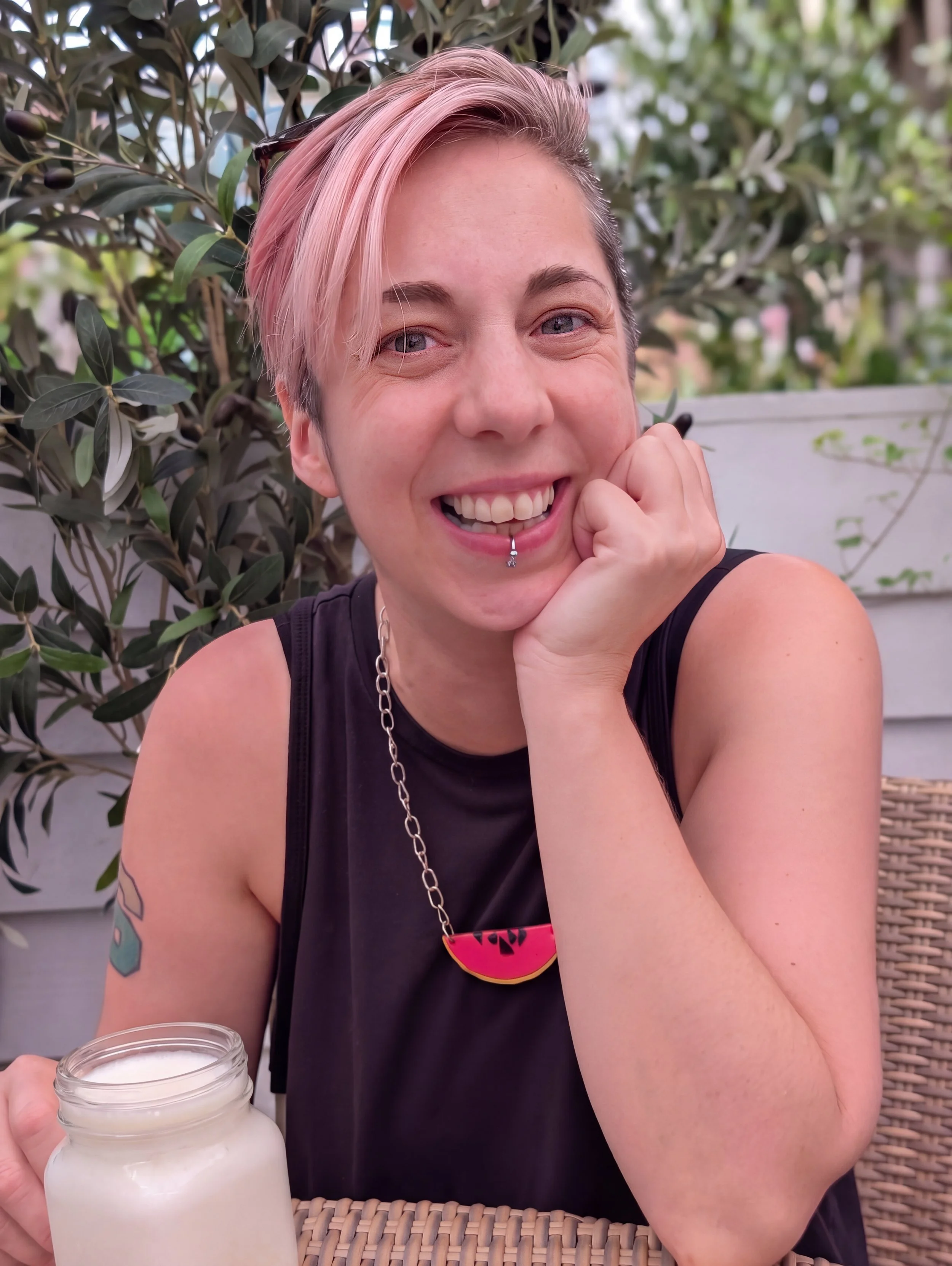 Mo Ford, a person with short pink hair and a lip piercing smiling while sitting outdoors, holding a cold coffee drink, with greenery in the background.
