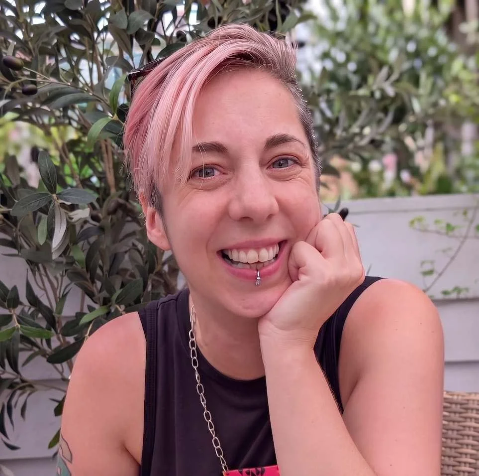 Mo Ford - a smiling person with short pink hair, wearing a black sleeveless shirt, and a silver chain necklace, sitting outdoors near green foliage.