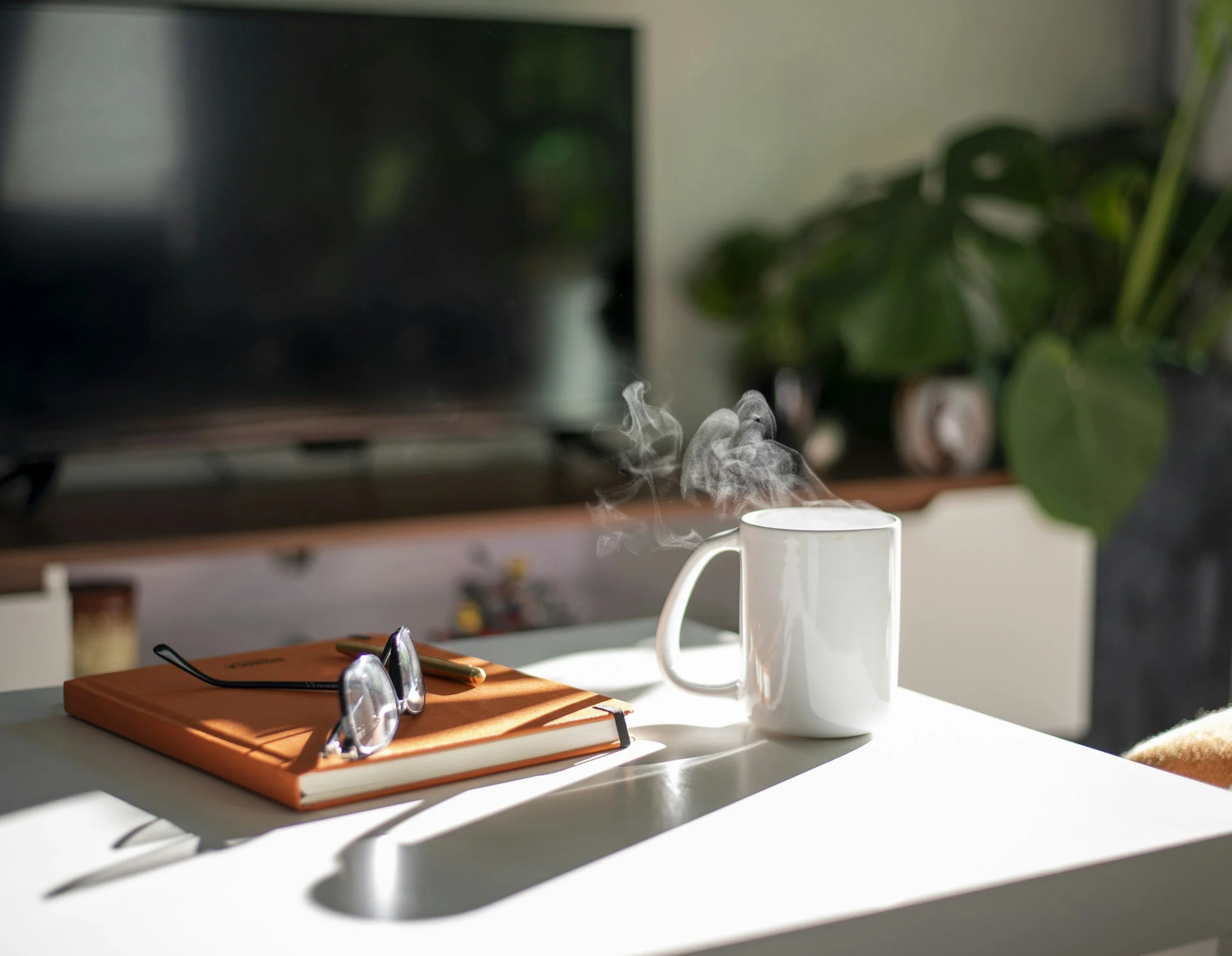 A white table with a closed orange notebook, a pair of eyeglasses, and a pen. Sunlight casts shadows on the table. In the background, there is a large monitor, a white mug with steam, and green plants.