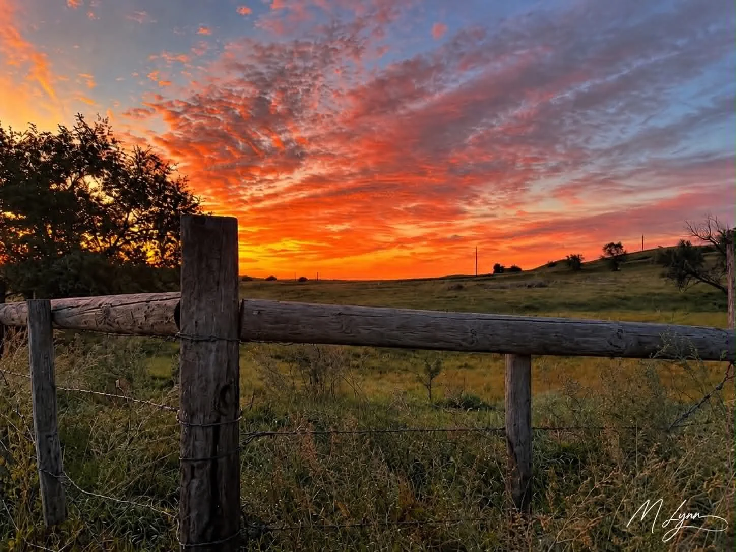 A rural landscape at sunset with a colorful sky filled with orange, pink, and purple clouds, a weathered wooden fence in the foreground, and rolling grassy hills with sparse trees in the background.