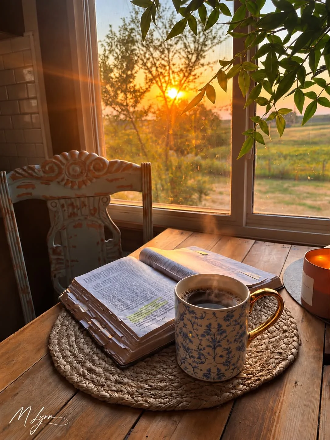 A cozy morning scene with a steaming cup of coffee on a wooden table, an open book, a candle, and a window showing a sunrise over a rural landscape with trees and fields, illuminated by warm sunlight.