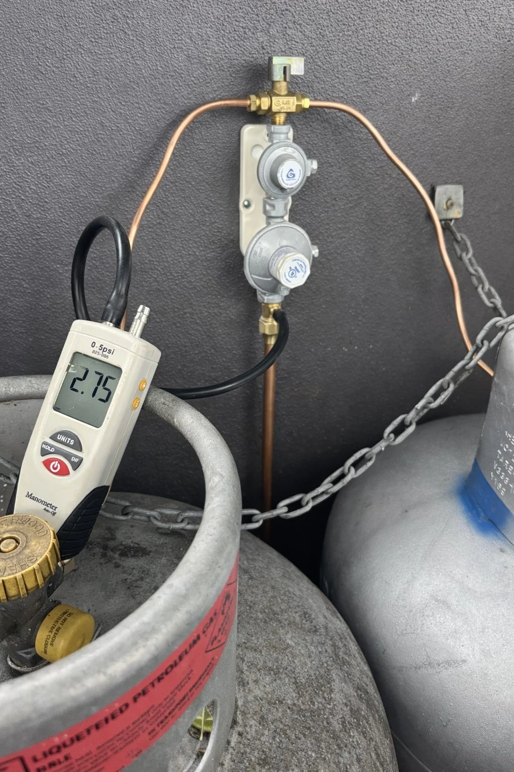 A propane tank setup with a pressure gauge reading 2.75 psi, connected to a regulator and copper piping, mounted on a gray wall with a chain securing the tank.