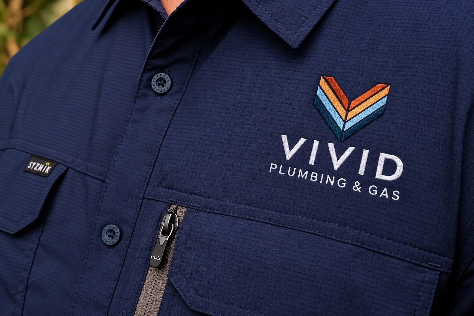 Close-up of a navy blue work jacket with a colorful embroidered logo and white text for Vivid Plumbing & Gas.