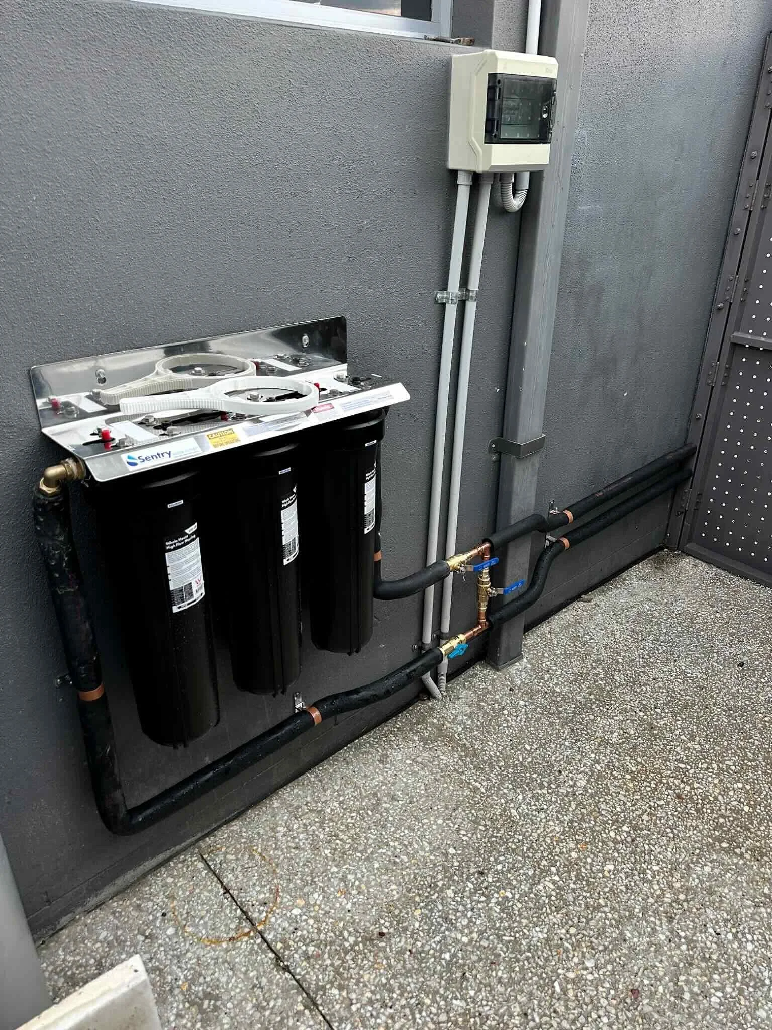 A utility panel with water filter system mounted on a gray wall outside, with pipes and valves below it on a textured concrete ground.