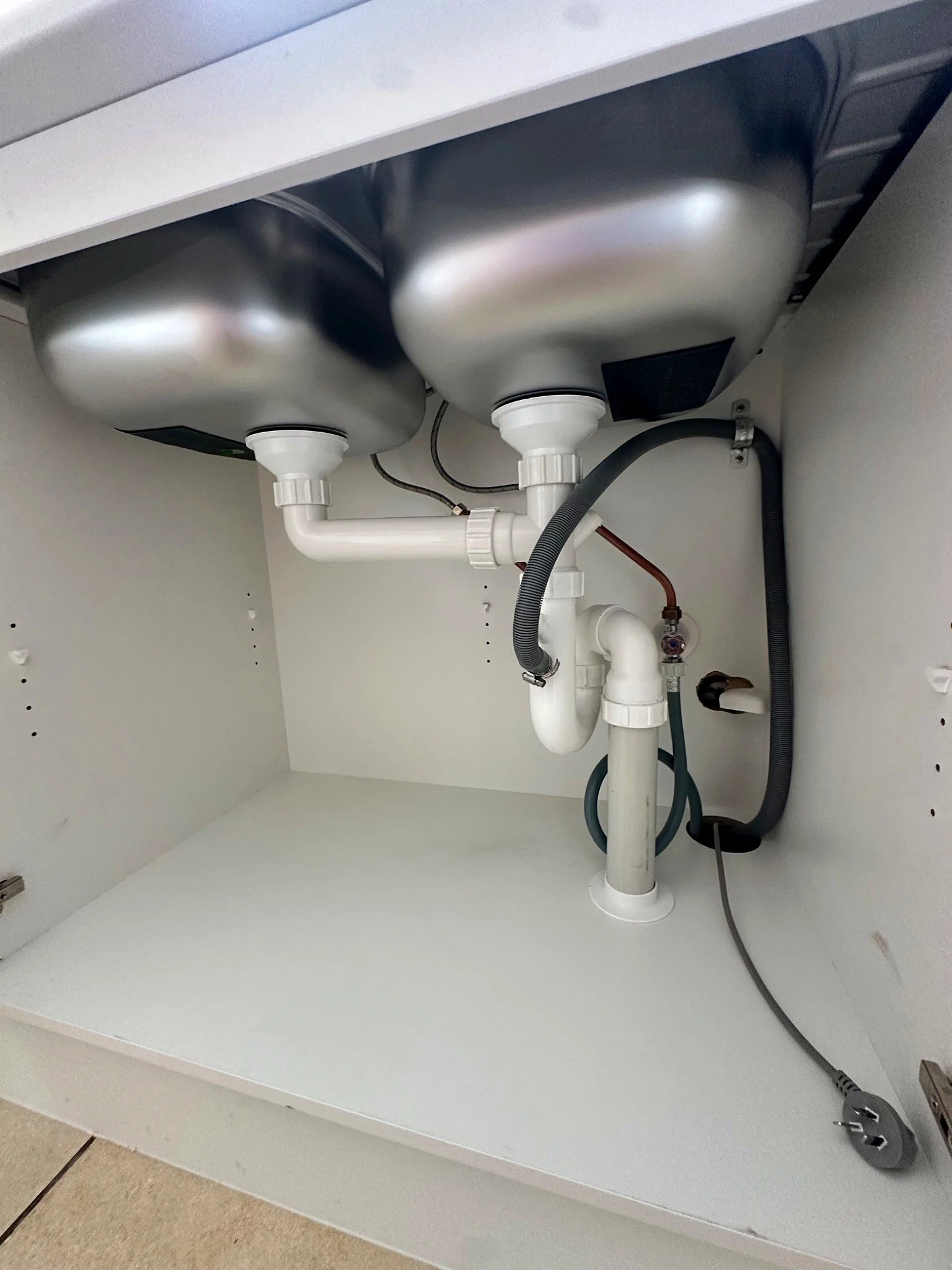 Underneath a kitchen sink showing plumbing pipes, hoses, and an electrical cord.