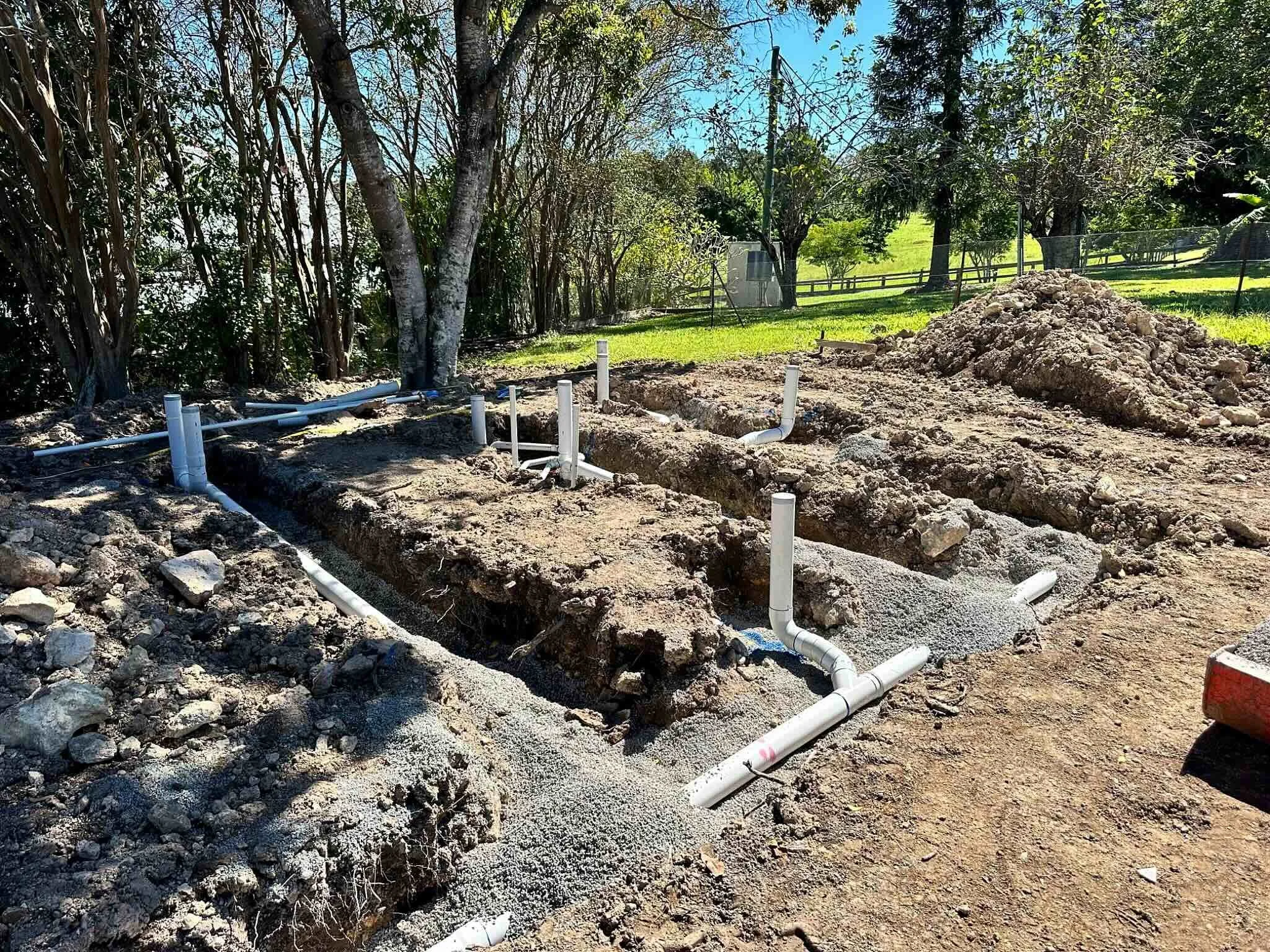 Construction site with exposed pipes and dug-up earth, surrounded by trees and grass, fencing, and a blue sky.