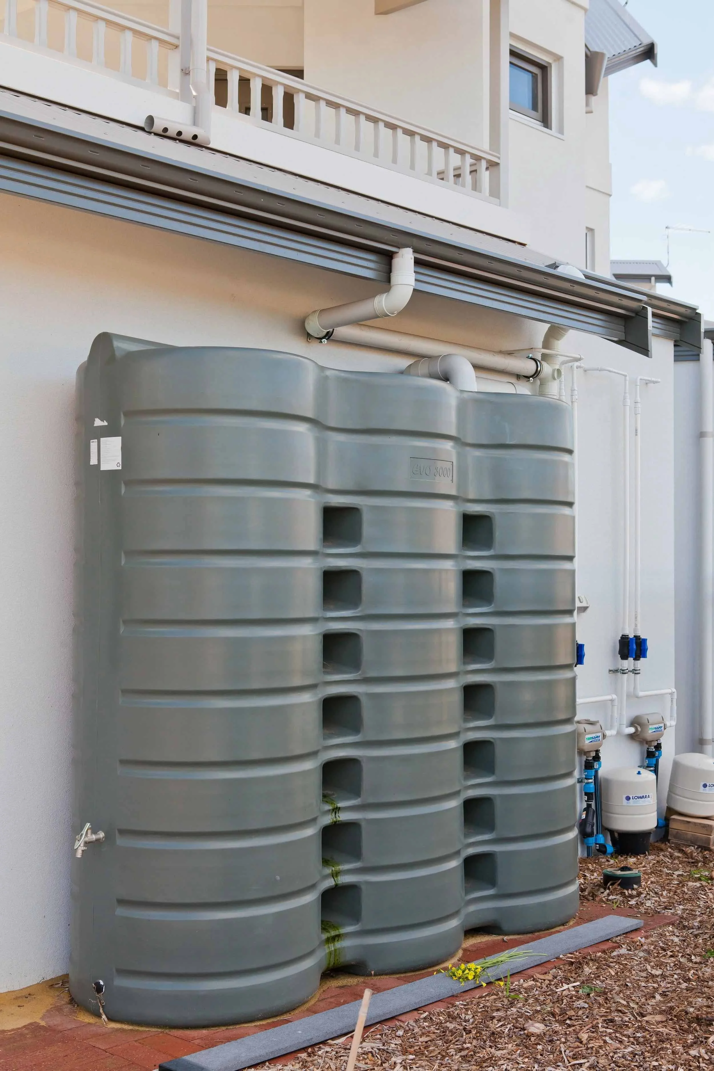 An outdoor space showing two large gray water storage tanks connected to a home's plumbing system. The tanks are mounted against a light-colored wall beneath a balcony with white railing. The area has some wood chips, a small patch of yellow flowers, and various pipes and meters for water management.