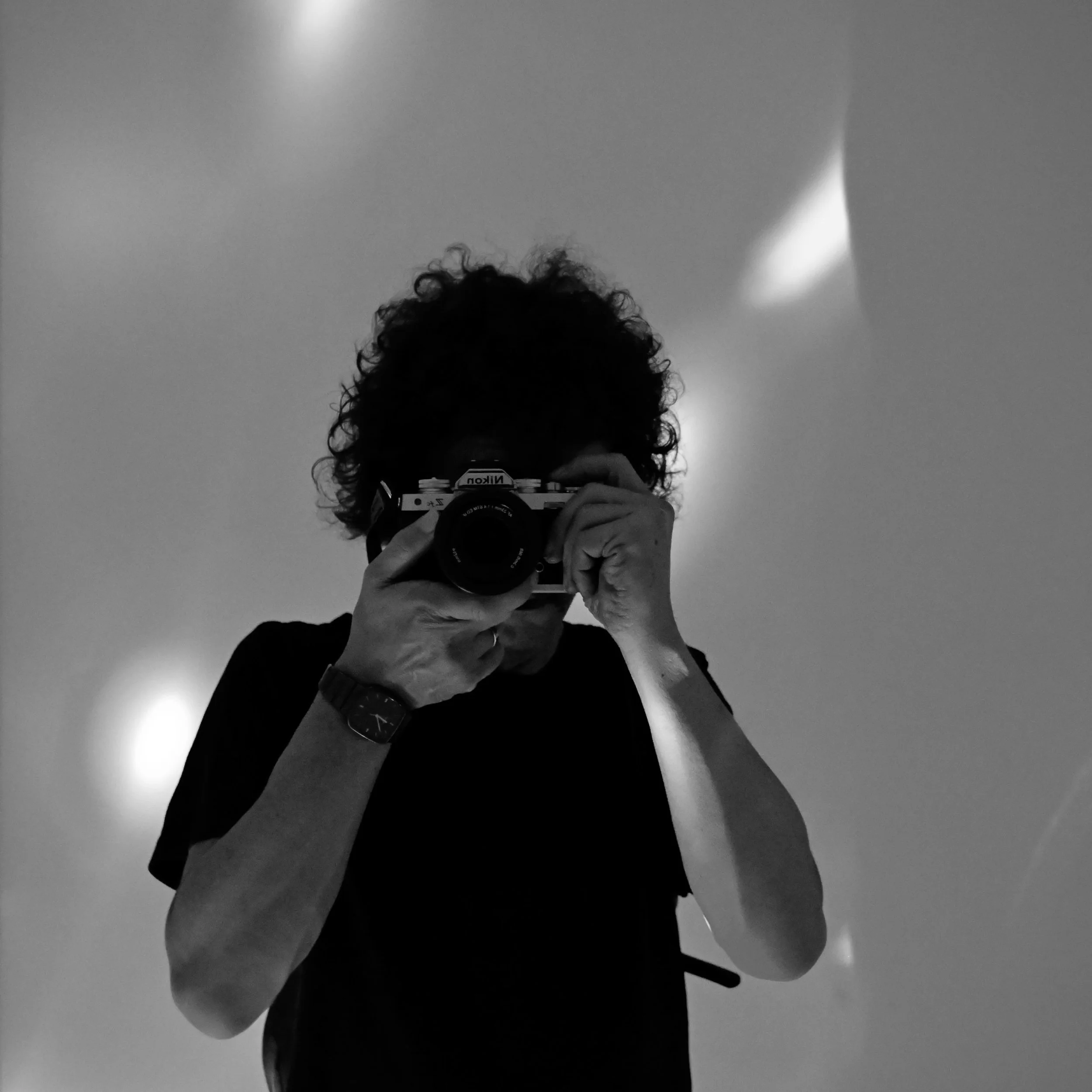 Schwarz-weiß-Foto eines Menschen, der ein Kamerasystem benutzt, um sich selbst zu fotografieren, mit einem Hintergrund mit grauer Wand und Lichtreflexen an der Decke.