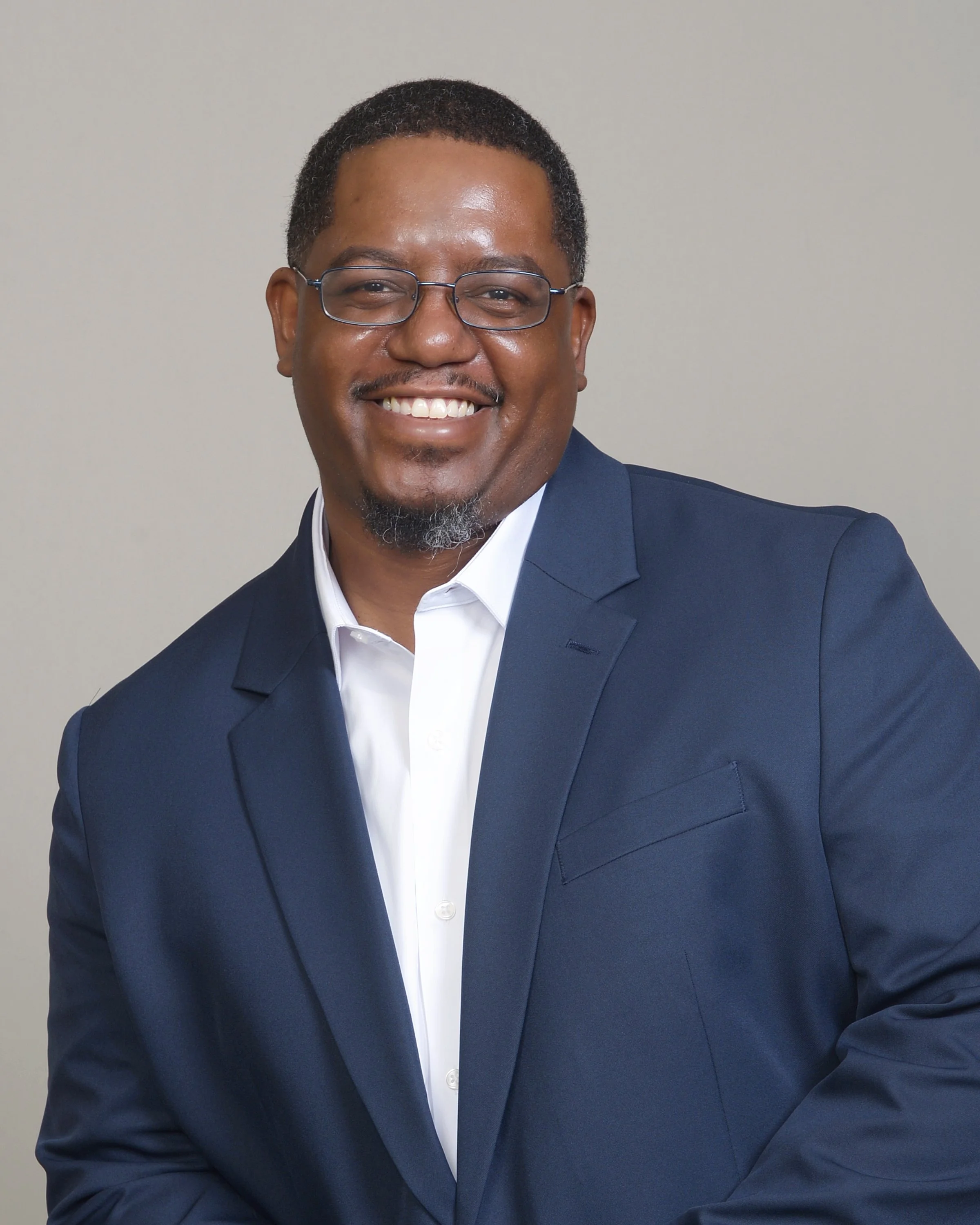 Portrait of a smiling man with glasses, wearing a navy blazer and white shirt, against a plain light gray background.