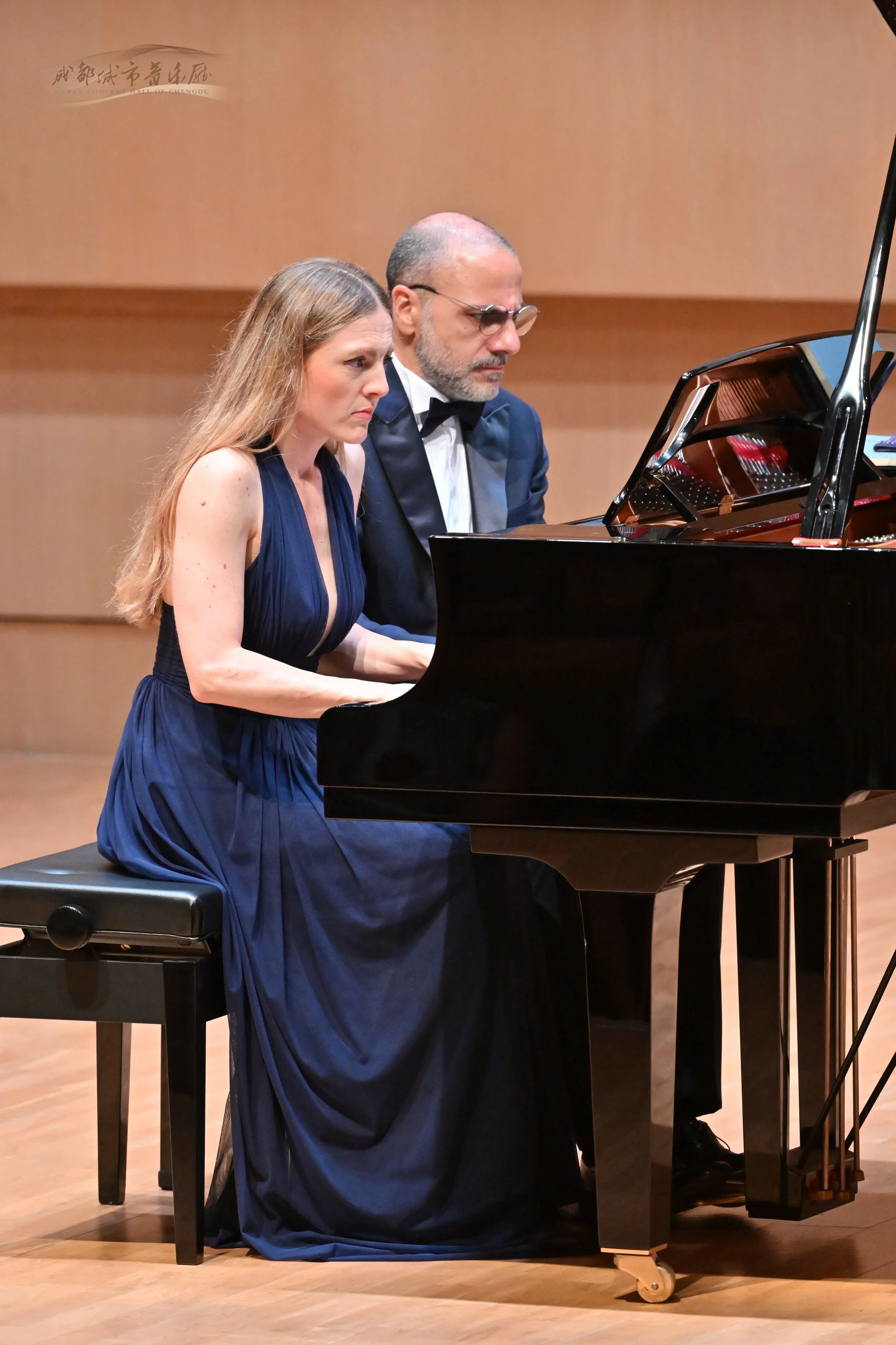 Due persone, una donna e un uomo, suonano un pianoforte a coda in un ambiente da concerto. La donna indossa un abito blu scuro, mentre l'uomo è in smoking con papillon. Entrambi sono concentrati sulla musica.